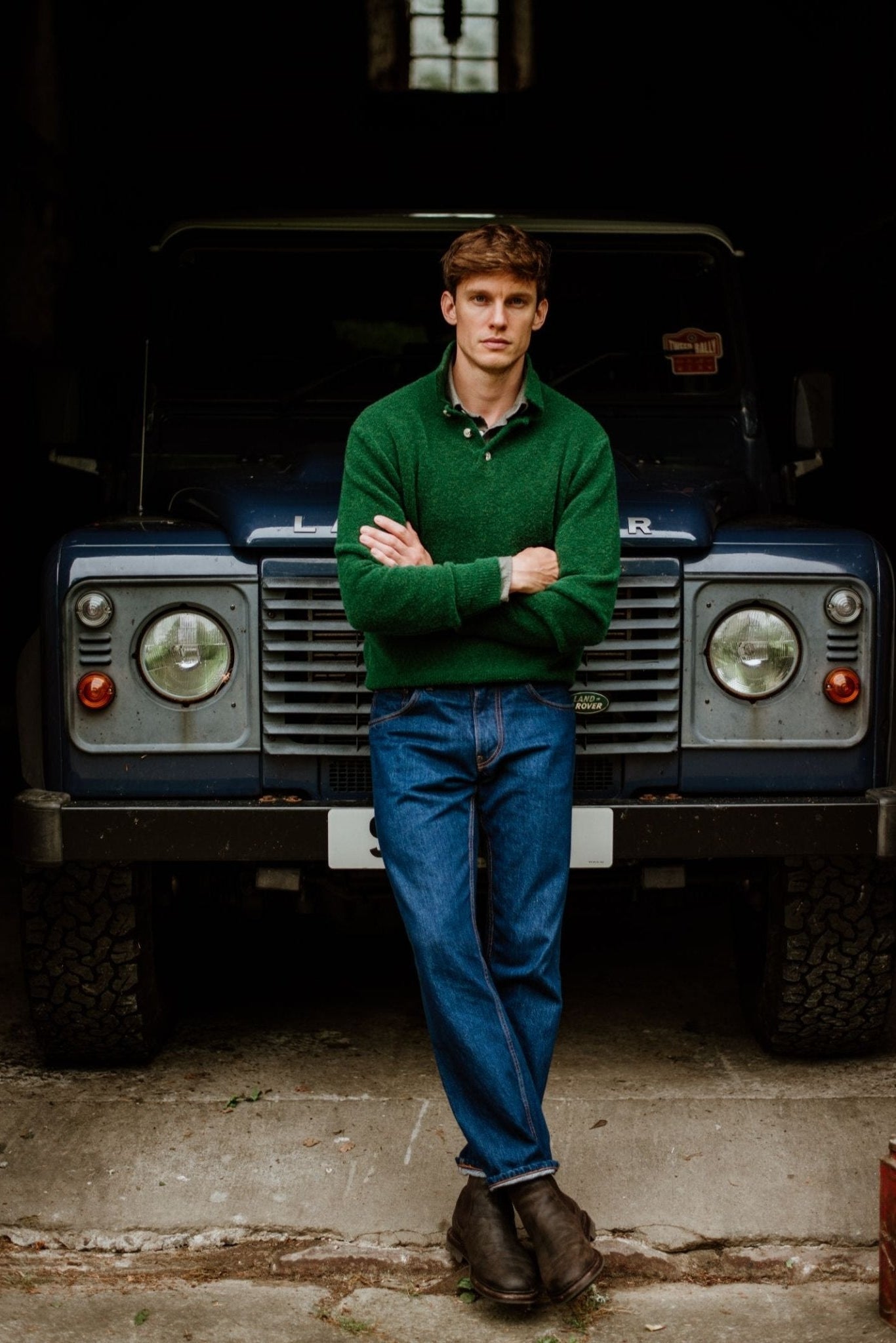 A man wearing a green Campbell's of Beauly Bouclé Polo Sweater and blue jeans stands with arms crossed in front of a dark blue Land Rover inside a dim garage.