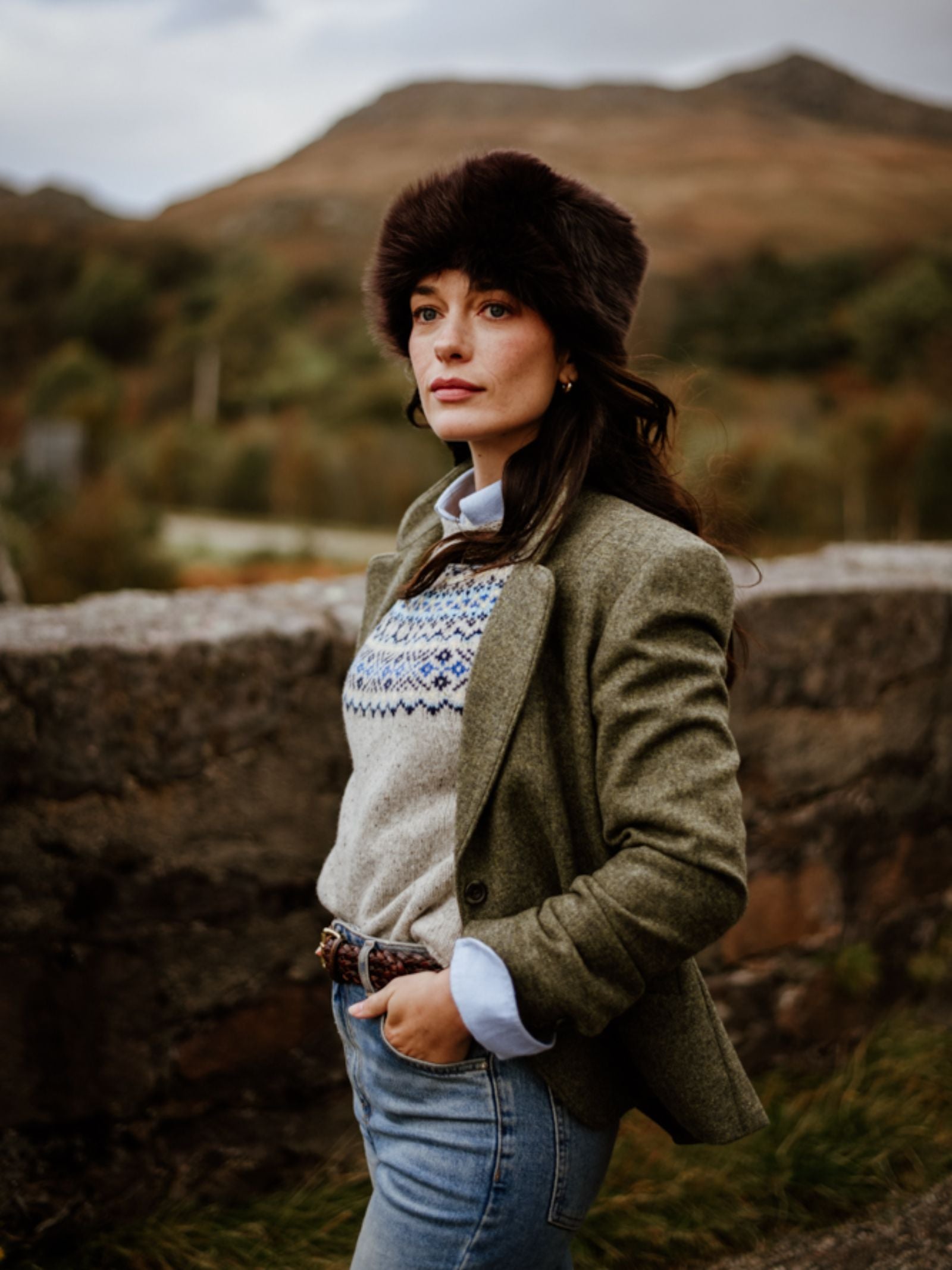 A woman stands outdoors by a stone wall, wearing Campbell's of Beauly House Jacket over a patterned sweater and jeans, topped with a furry hat. Behind her, blurred hills and trees appear under a cloudy sky.
