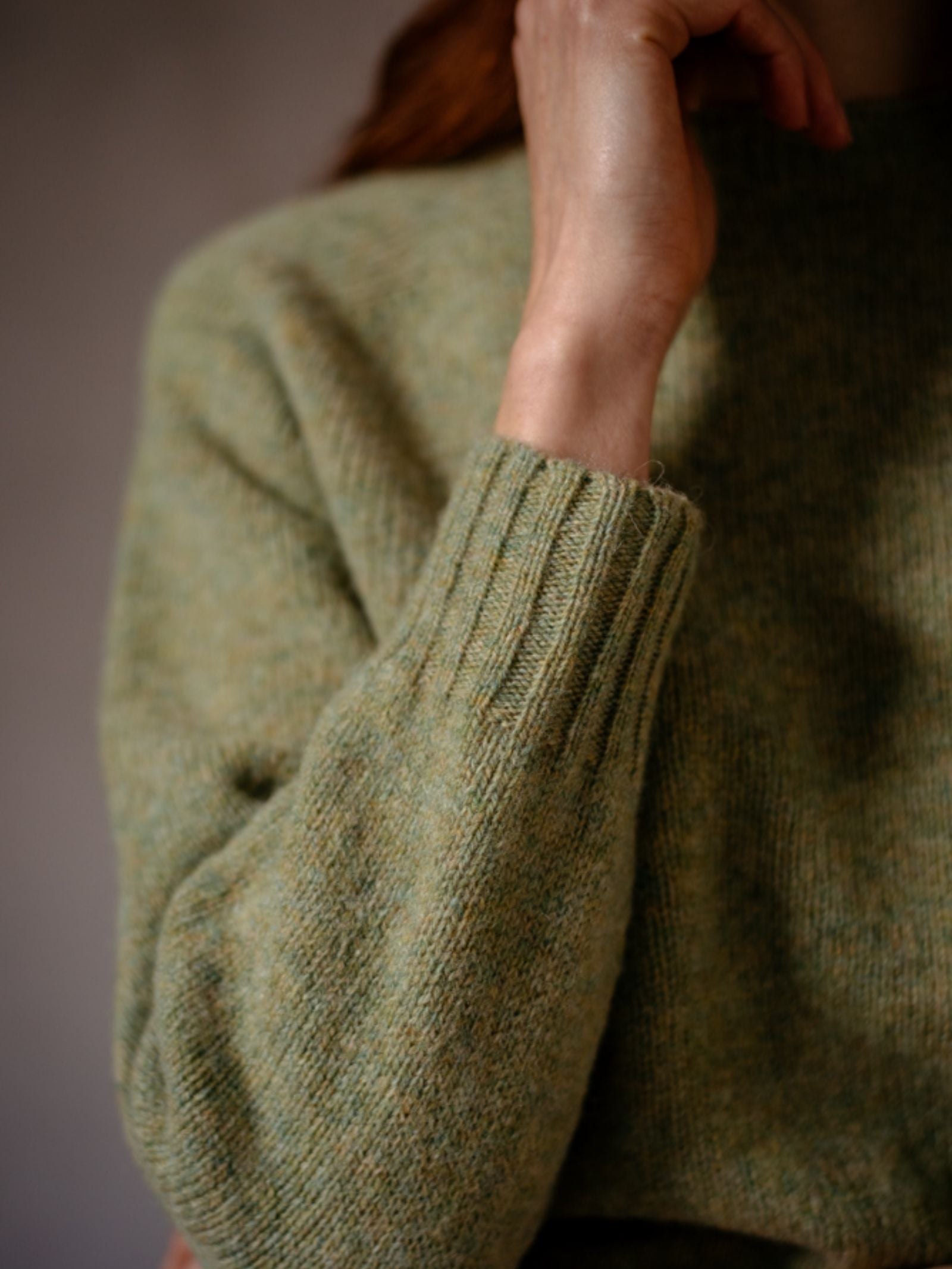 A person wears the Campbells of Beauly Cropped Shetland Jumper in textured light green Shetland lambswool, shown from shoulders to mid-torso with a hand raised near their chin against a softly blurred, warm background.
