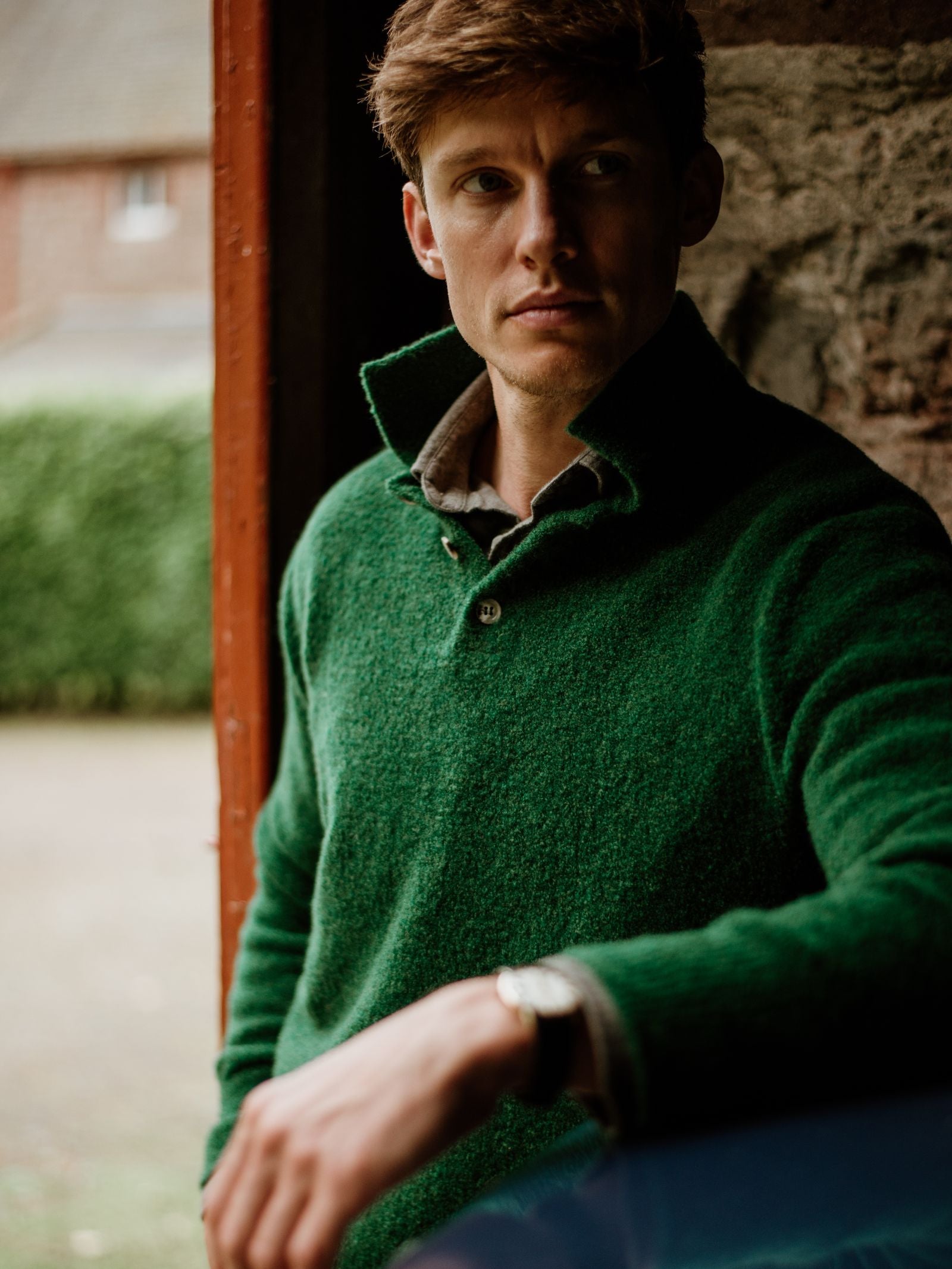 A man with light brown hair wears the Campbell's of Beauly Bouclé Polo Sweater over a shirt, standing indoors by a stone wall as soft natural light highlights his thoughtful expression.