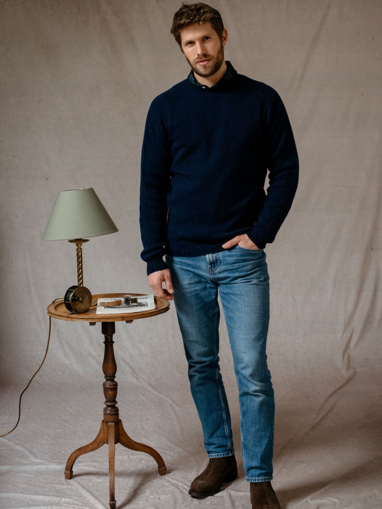 A man with short brown hair and a beard stands indoors by a small round wooden table with a green lamp, books, and glasses. He wears the Campbell's of Beauly Shetland Jumper in navy, light blue jeans, and brown boots against a plain backdrop.