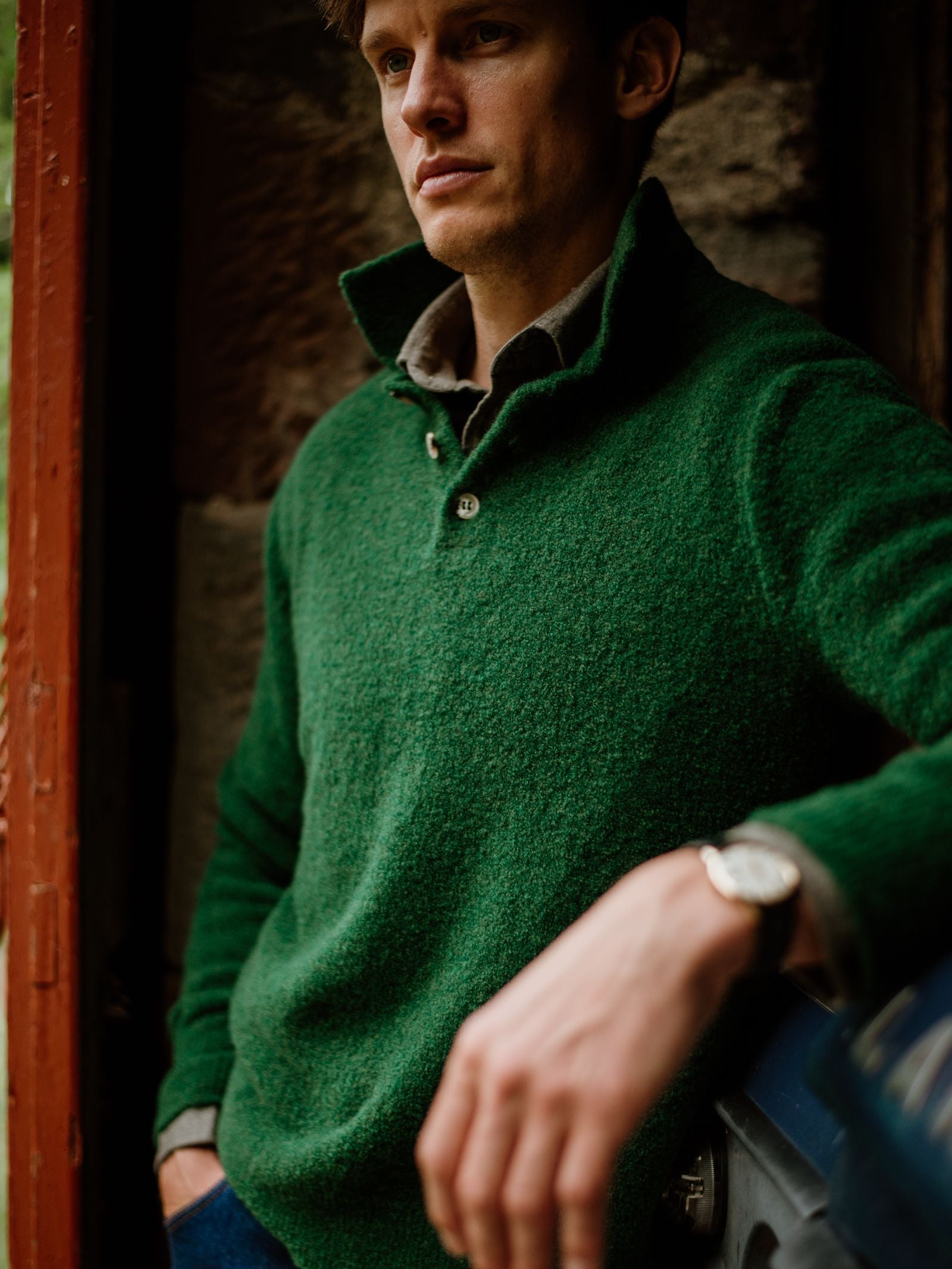 A man stands indoors, leaning against a surface, wearing the Campbell's of Beauly Bouclé Polo Sweater with the collar up. His face is partially visible, left hand in focus, and he sports a wristwatch.