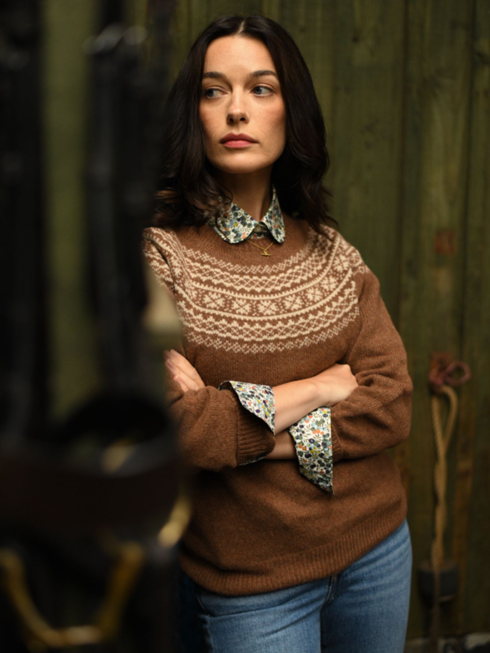 A woman with dark hair stands, arms crossed, wearing the Campbell's of Beauly Two-Colour Fairisle Yoke Jumper over a floral shirt and blue jeans, gazing to the side against a rustic green wooden backdrop.