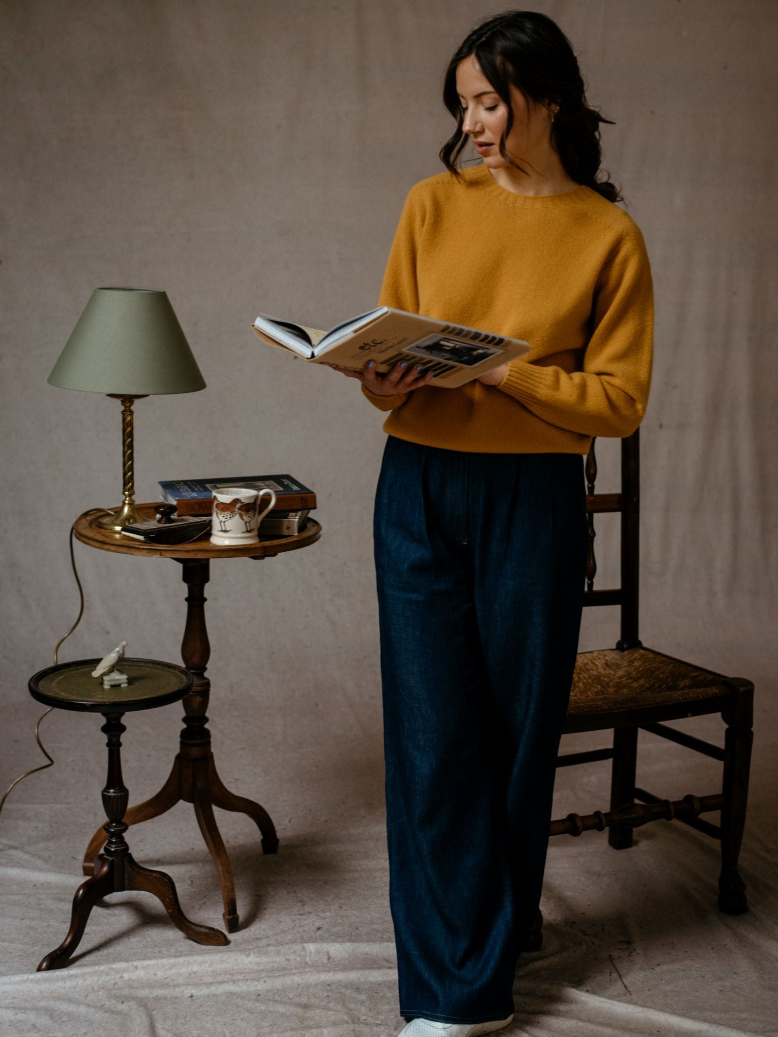 A woman in a Campbells of Beauly Geelong Lambswool Crew Neck Jumper and wide-leg jeans stands by a wooden chair, reading. Nearby is a small table with books, a lamp, mug, and figurine. The simple background highlights the Scottish craftsmanship.