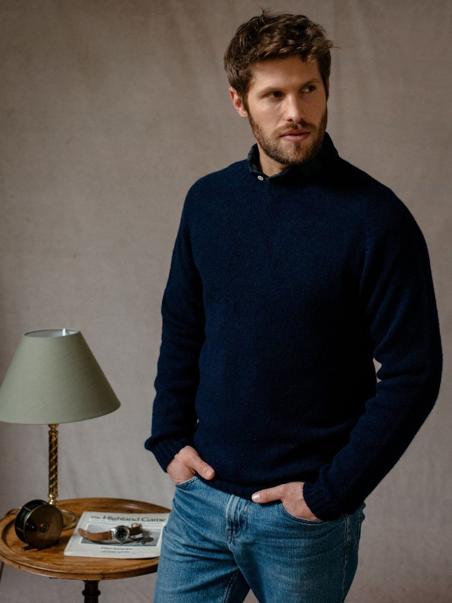 A man with short brown hair and a beard wears a dark blue Shetland Jumper by Campbells of Beauly and jeans, standing with his hands in his pockets. He looks to the side beside a small round table holding a lamp, camera, and magazines.