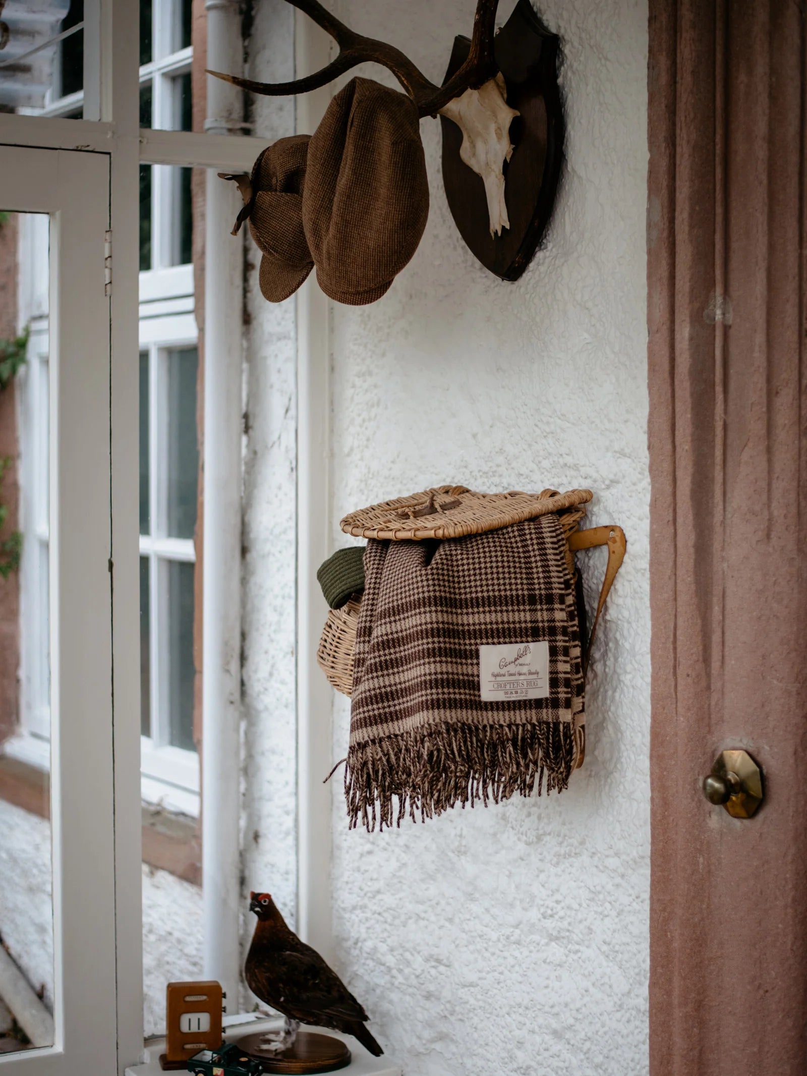 A cozy entryway with hats on antlers, a country look highlighted by the Campbells of Beauly Crofters Blanket in brown houndstooth, a woven basket, and door handle. A decorative bird sits on a small table under the window.