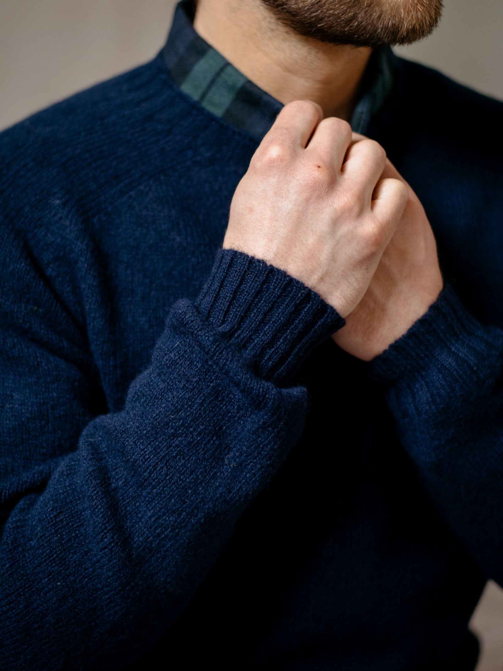 A man adjusts the collar of his green and navy plaid shirt, layered under a Campbells of Beauly Shetland Jumper. Only his lower face and upper torso are shown, highlighting this classic heritage knitwear style.