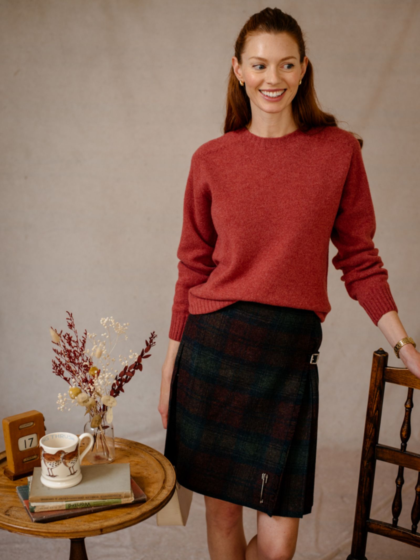 A woman in a red Campbells of Beauly Geelong Lambswool Crew Neck Jumper and a plaid skirt stands by a wooden chair and small table with books, a ceramic mug, dried flowers, and a calendar showing 17. She smiles and looks to the side.