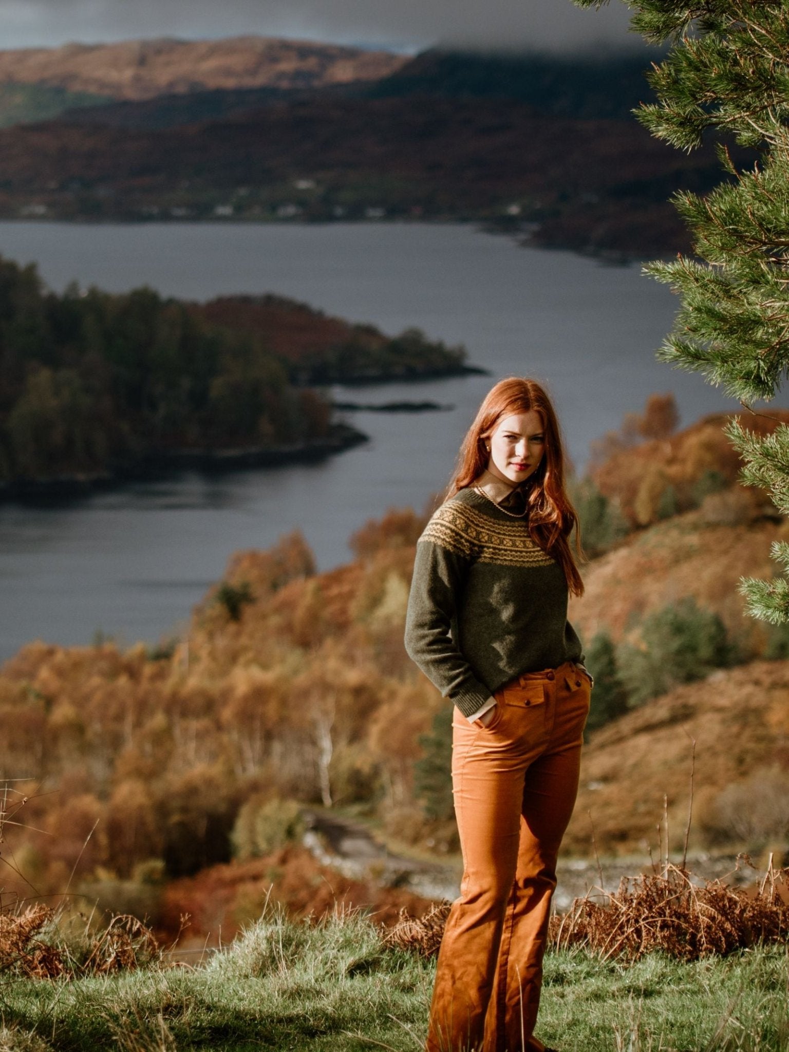 A woman with long red hair, wearing Campbells of Beauly’s Two-Colour Fairisle Yoke Jumper and orange pants, stands on a grassy hillside overlooking a lake with autumn trees and distant hills in the background.