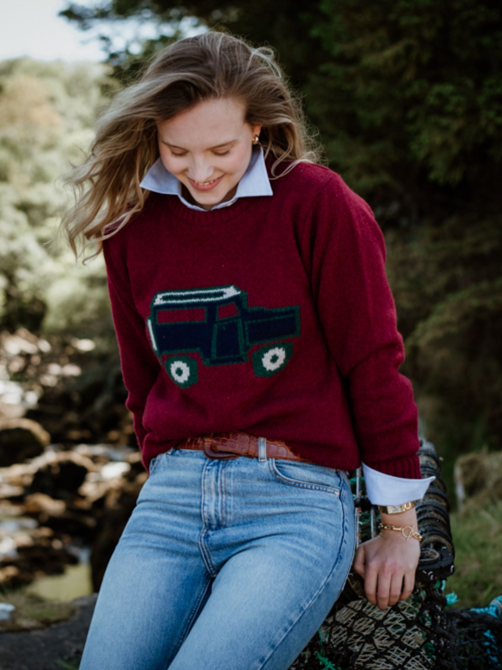 A woman with light brown hair, wearing the Campbells of Beauly Land Rover Defender Jumper in claret over a collared shirt and blue jeans, sits outdoors smiling and looking down, with trees and rocks in the background.