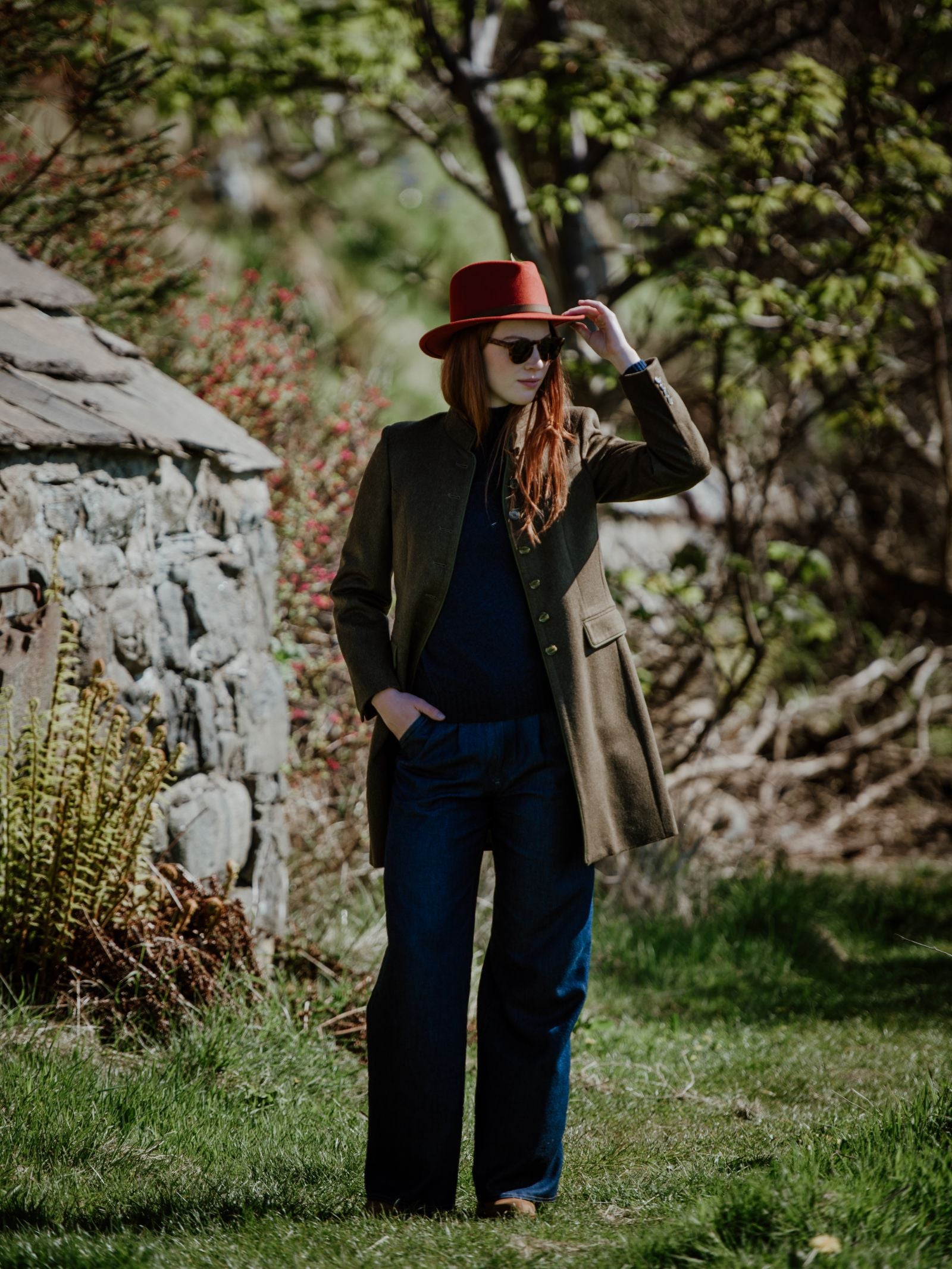 A person wearing a red hat, sunglasses, and the Campbells of Beauly Loden Green Millie Coat made from Austrian loden wool stands on grass by a stone building amid greenery and trees.