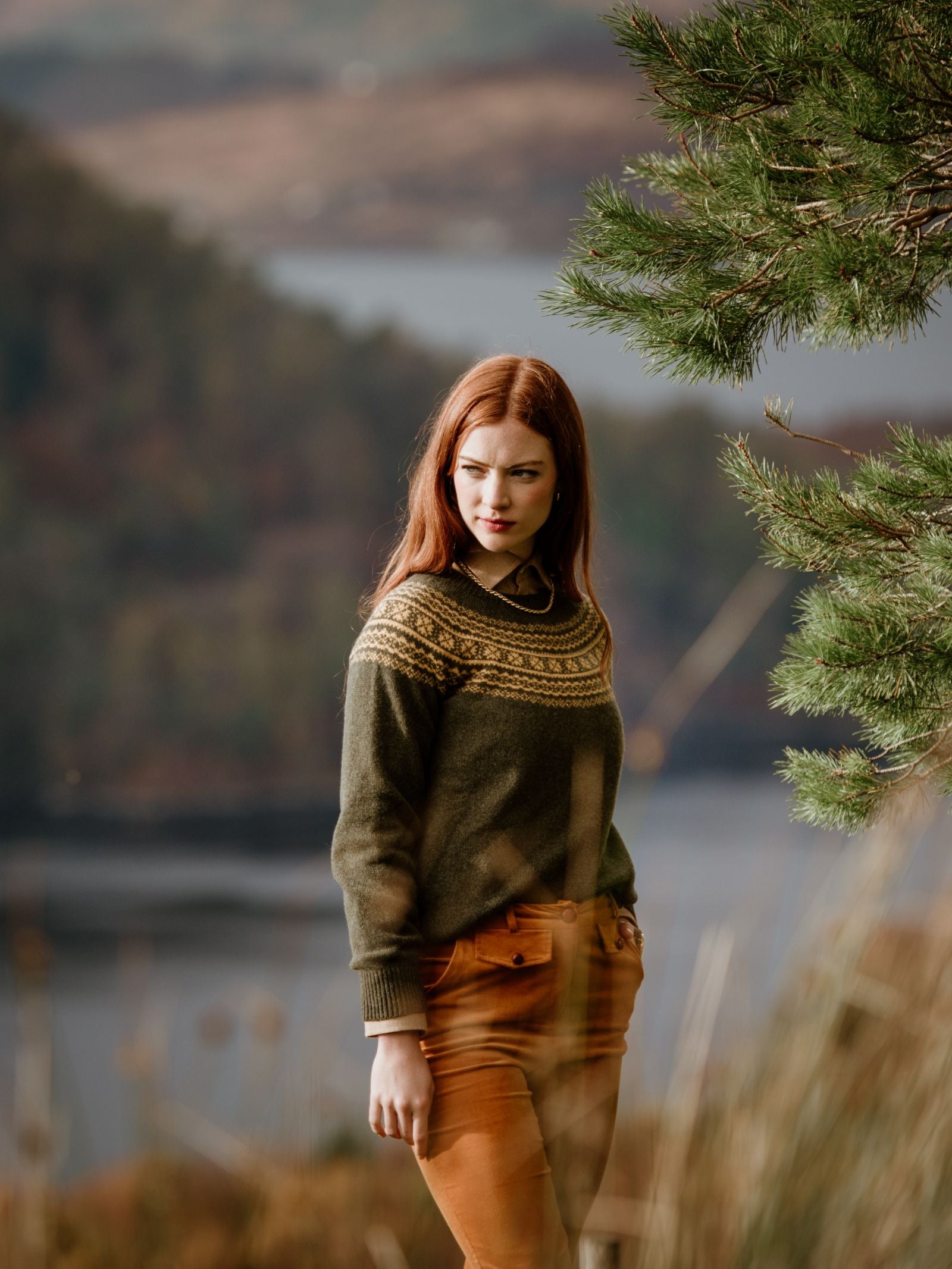 A woman with long red hair wears a Campbells of Beauly Two-Colour Fairisle Yoke Jumper and tan pants, standing outdoors by a pine tree with blurred hills and water in the background.