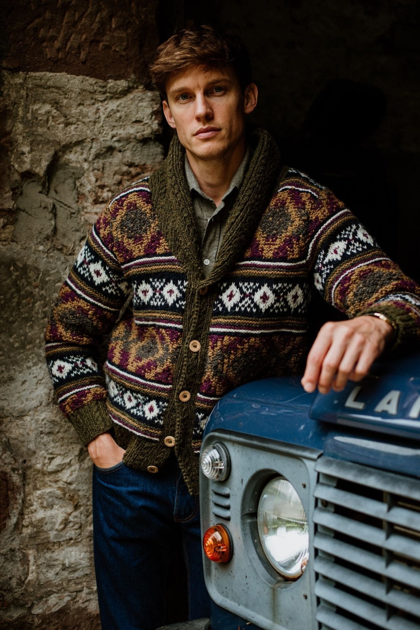 A man in a Campbell's of Beauly Navajo Shawl Collar Cardigan and jeans leans against a vintage blue Land Rover by a stone wall, looking seriously at the camera.