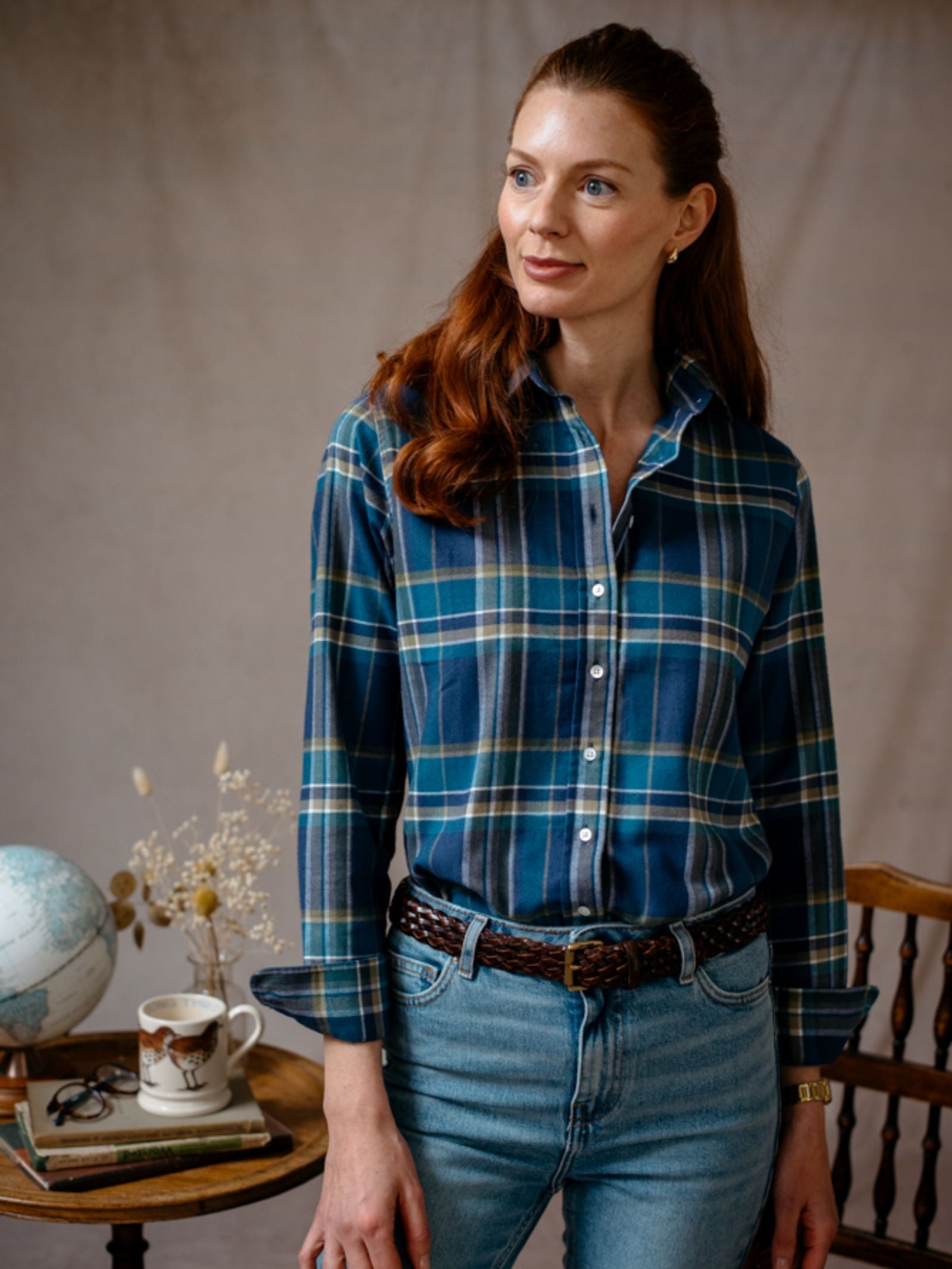 A woman with long red hair, wearing Campbells of Beauly Brushed Cotton Shirt in blue plaid and light jeans, stands indoors by a table with books, a mug, a globe, and dried flowers. She looks calmly to the side.