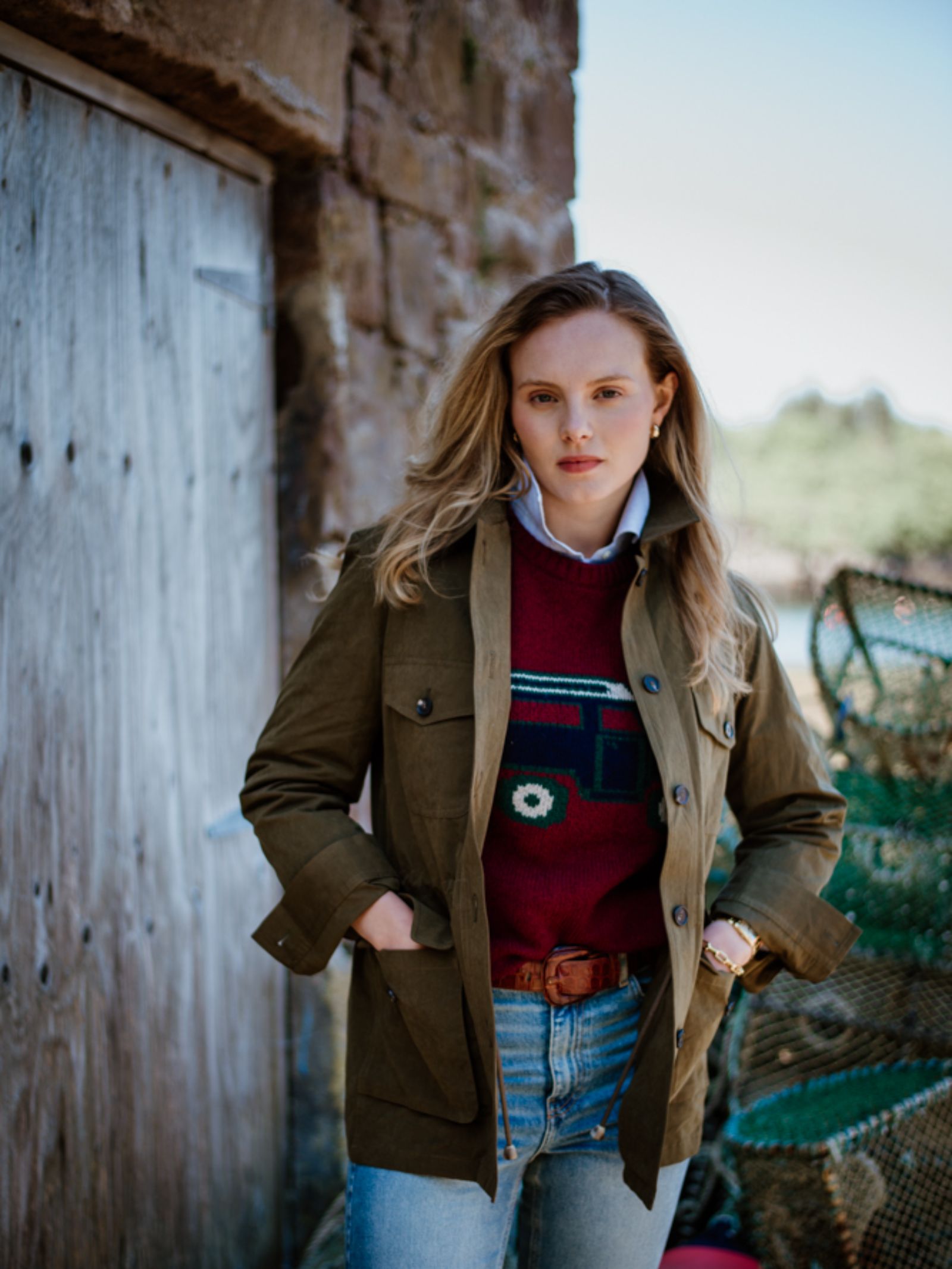 A woman with long blond hair stands outside by a wooden door and stone wall, wearing a Campbells of Beauly Land Rover Defender Jumper over a white shirt, green jacket, jeans, and brown belt, hands tucked in her jacket pockets.