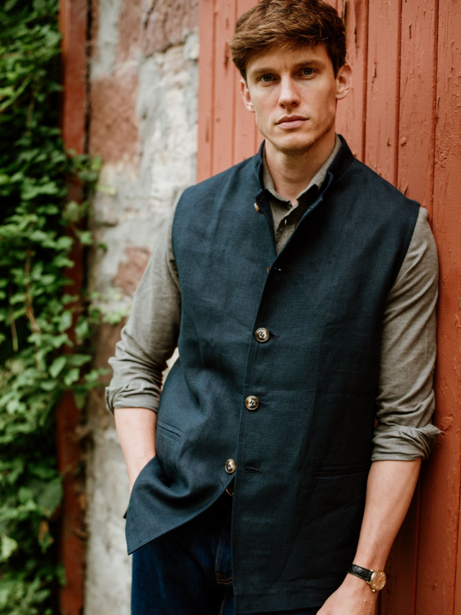 A man with light brown hair stands against a red wooden wall in a Navy Linen Nehru Gilet by Campbell’s of Beauly over a gray shirt, one hand in his pocket, looking at the camera. Green plants can be seen nearby.