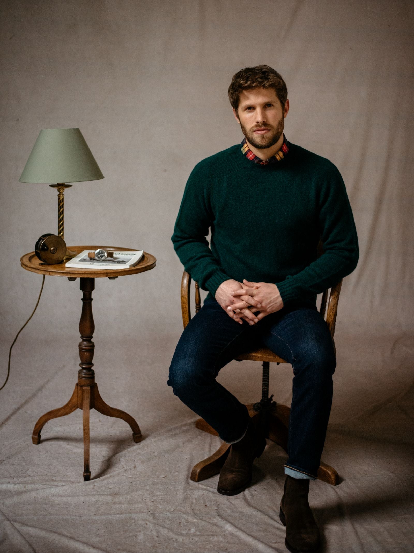 A man with brown hair and a beard sits on a wooden chair, wearing a dark green Campbells of Beauly Shetland Jumper. Beside him is a small wooden table with a lamp, book, and glasses; draped cloth forms the backdrop.