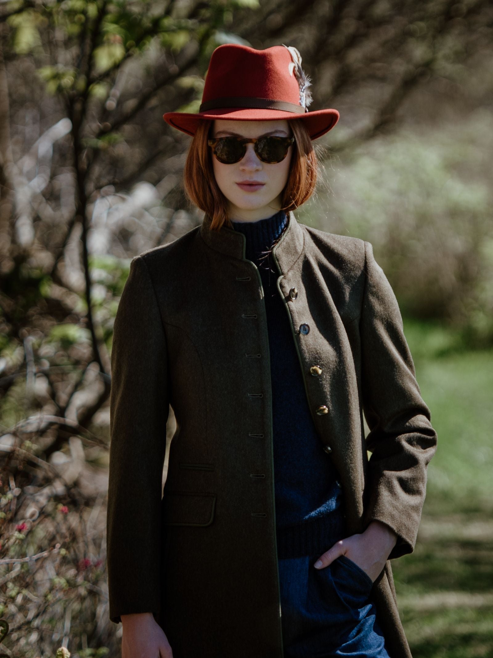 A person stands outdoors in greenery wearing Campbells of Beauly Loden Green Millie Coat, a red feathered hat, round sunglasses, and a blue turtleneck, with one hand in their pocket.