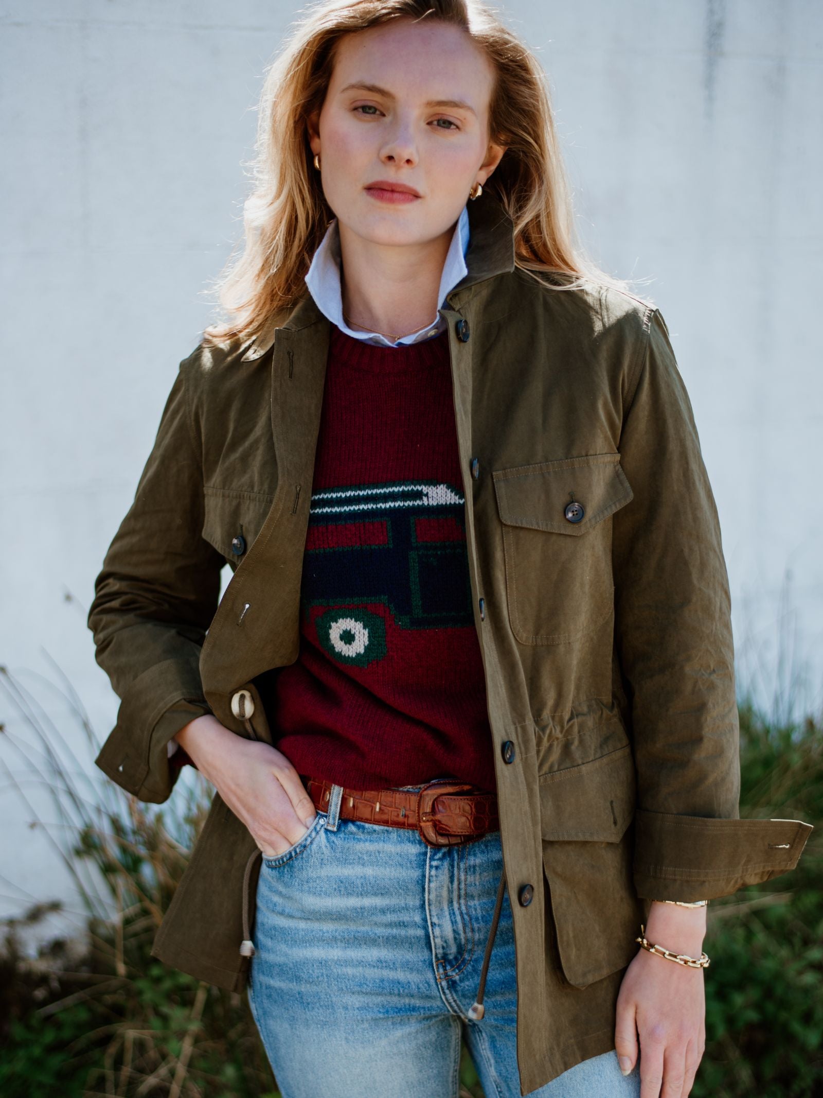 A blonde woman wears the Campbell's of Beauly Auld Stock Aigas Field Jacket, a green water-repellent peach skin jacket, over a maroon car-graphic sweater, blue collared shirt, light blue jeans, and brown belt, outdoors by a white wall.