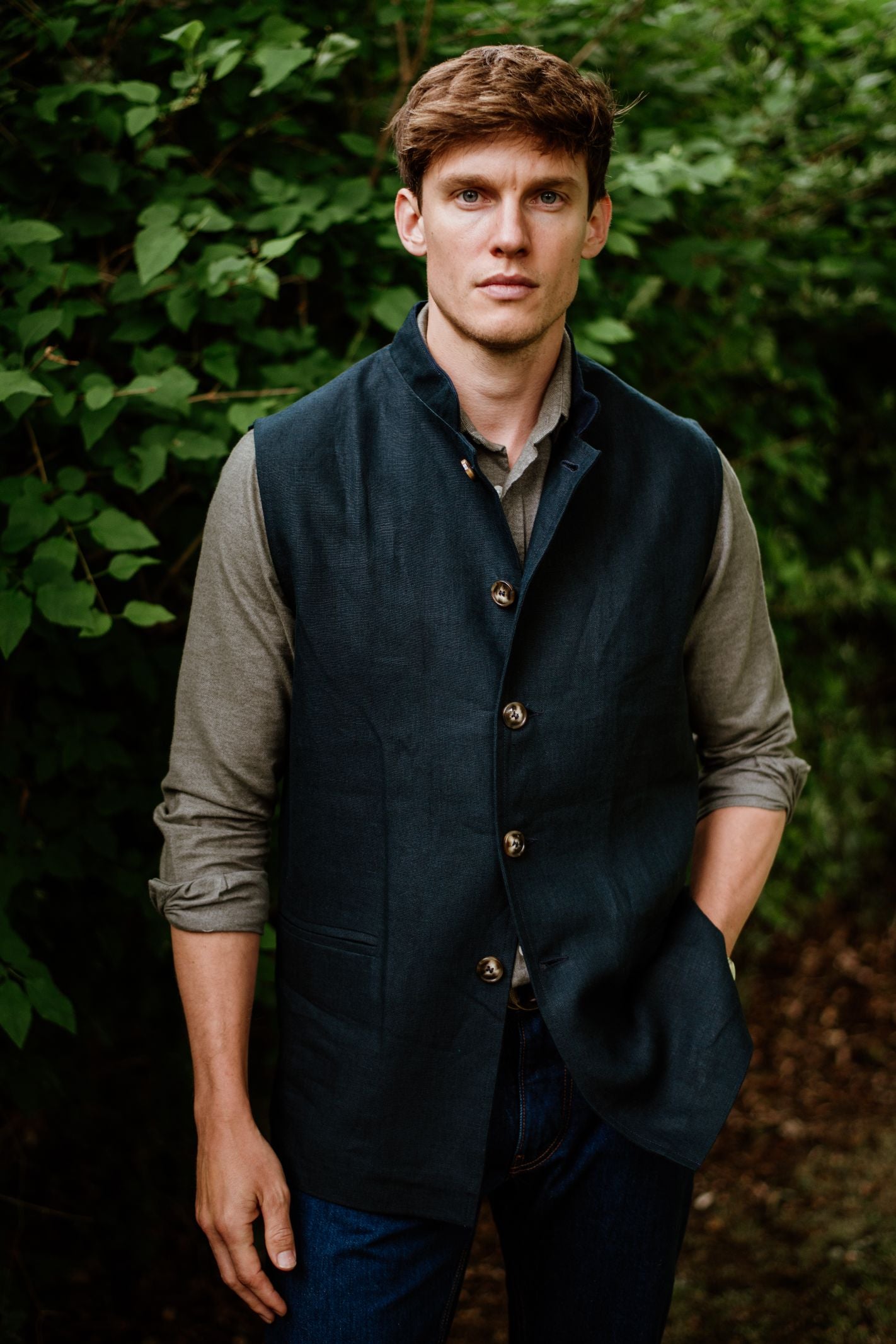 A young man with short brown hair stands outdoors before green foliage, wearing the Campbell’s of Beauly Linen Nehru Gilet over a light long-sleeve shirt and blue jeans—a perfect summer layer, with one hand in his pocket.