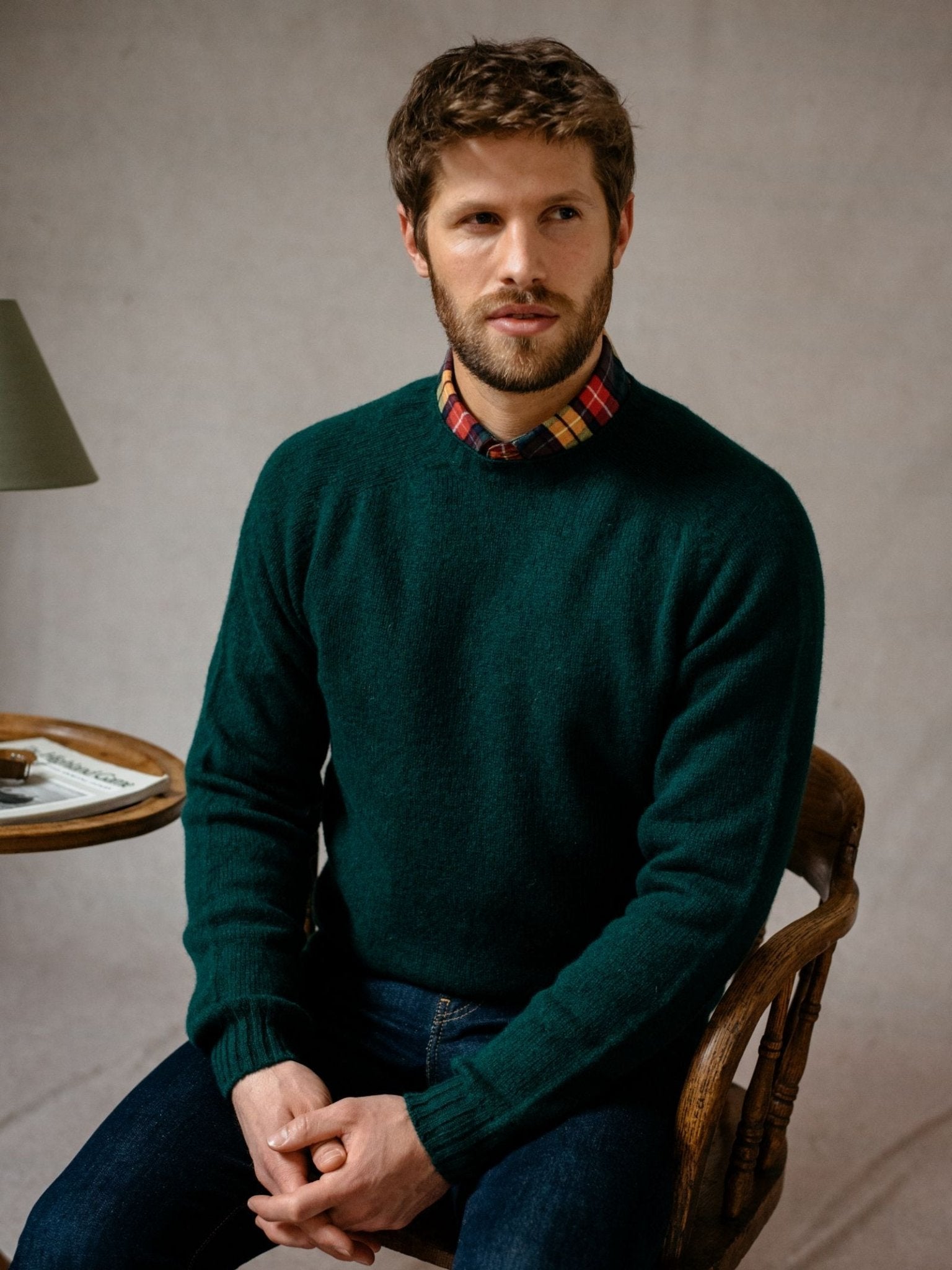 A man with short brown hair and a beard sits on a wooden chair, wearing the Campbells of Beauly Shetland Jumper in heritage-inspired colours over a red and yellow plaid shirt. A table with a lamp and newspaper is beside him, his hands folded in his lap.