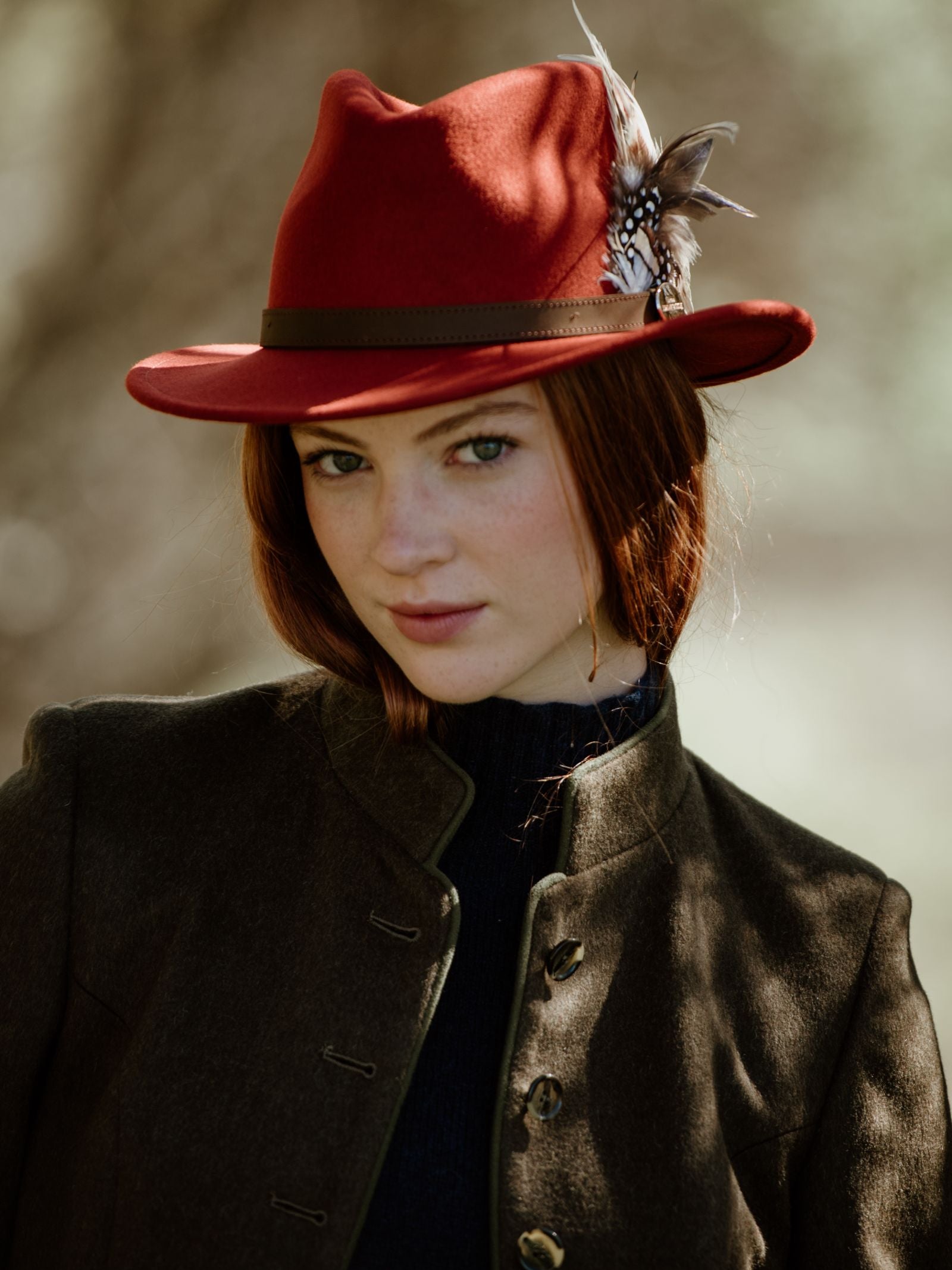 A young woman with red hair wears the Hicks & Brown Gamebird Fedora, featuring a feather wrap, and a dark buttoned jacket. She gazes at the camera outdoors in soft, natural light with a blurred background.