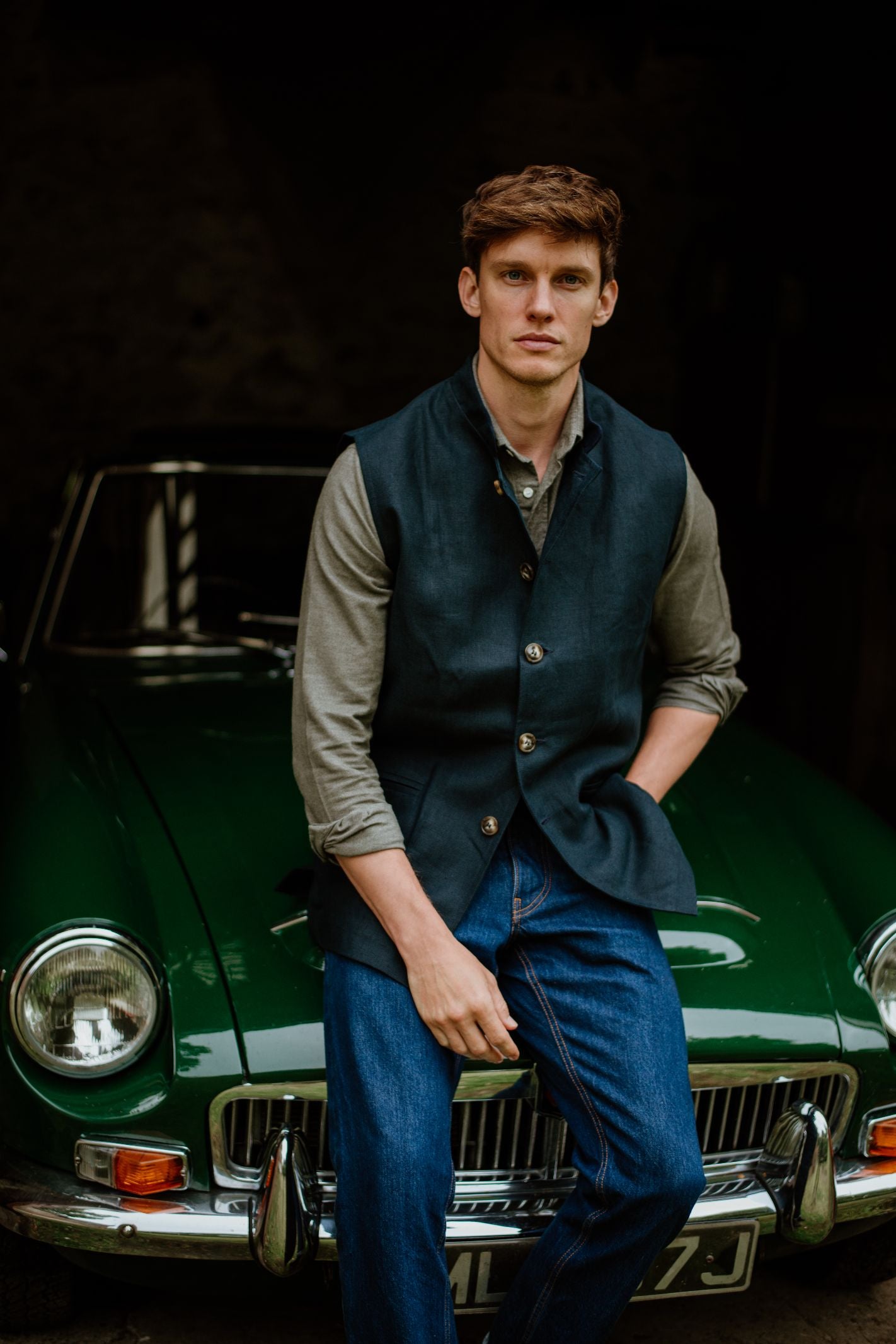 A man with short brown hair, dressed in a grey shirt, blue jeans, and a Campbells of Beauly Linen Nehru Gilet, leans casually against a classic green car and gazes towards the camera.