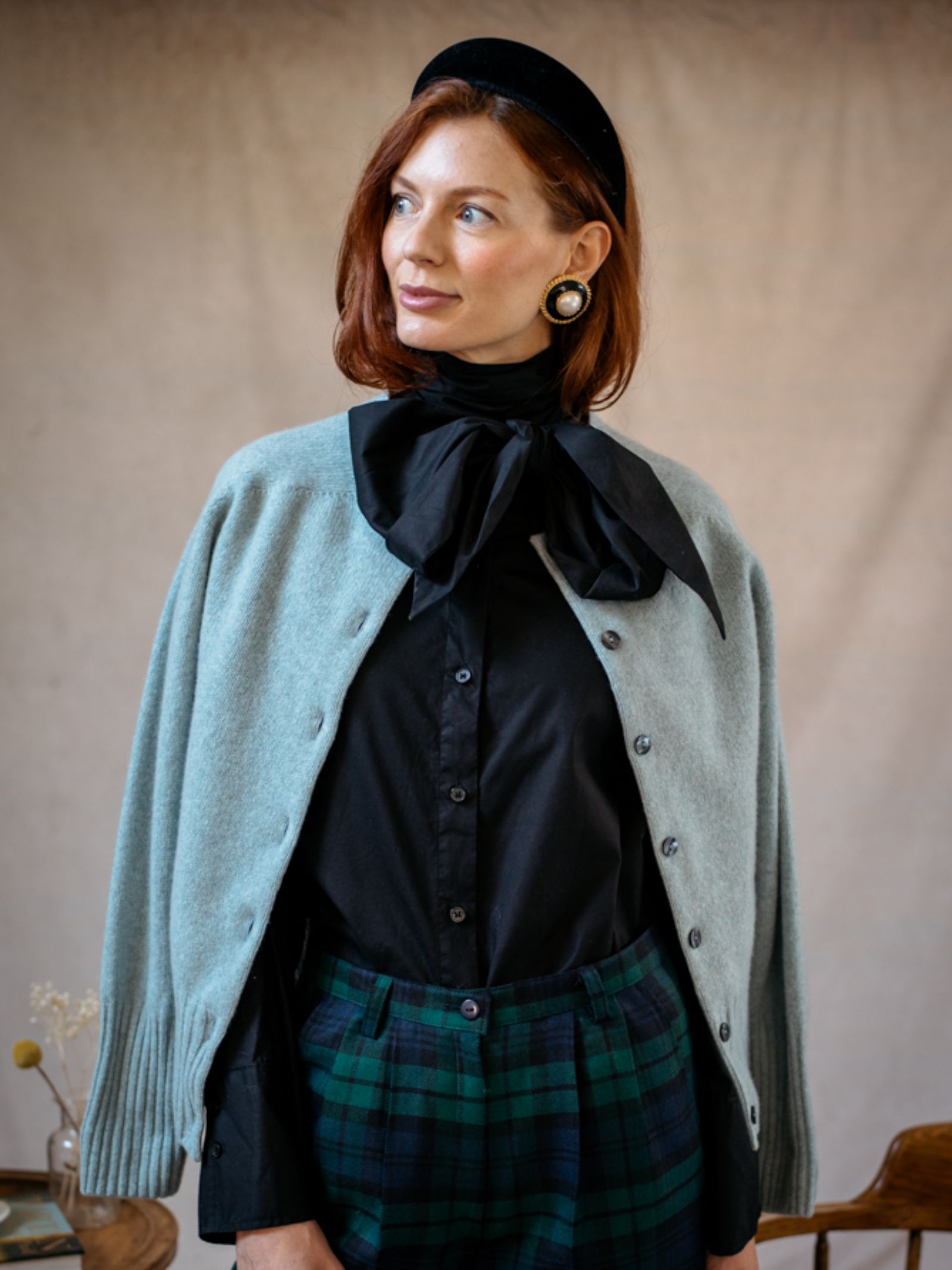 A woman with red hair wears a black blouse with a large bow, green plaid pants, a black hat, round earrings, and the Campbells of Beauly Crew Cardigan draped over her shoulders, standing indoors against a neutral background.