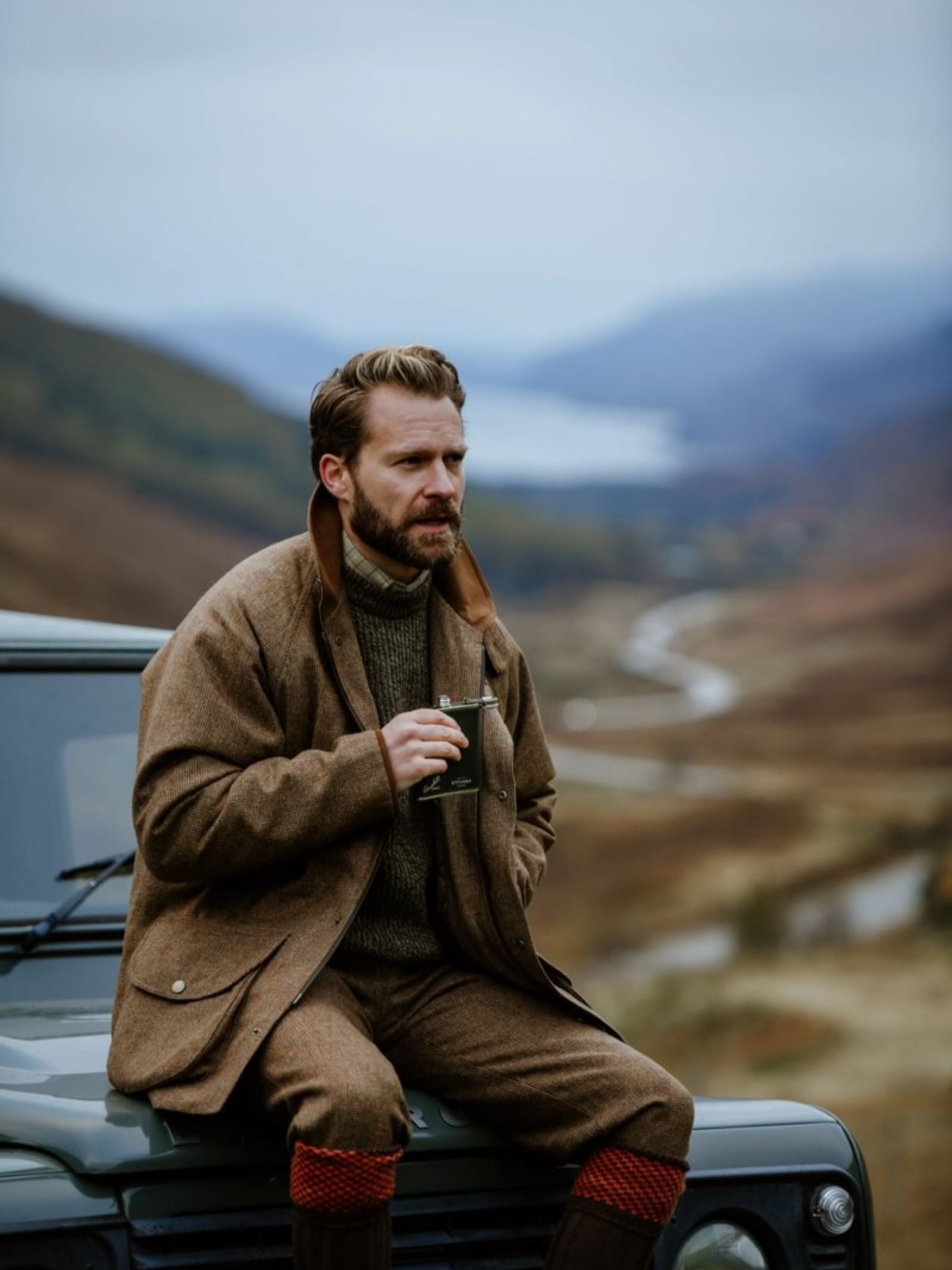 A man in brown tweed sits on a green car, holding a 6oz Forest Green Hip Flask by Campbell's of Beauly x Ettinger, with hills, a winding river, and cloudy skies in the background.