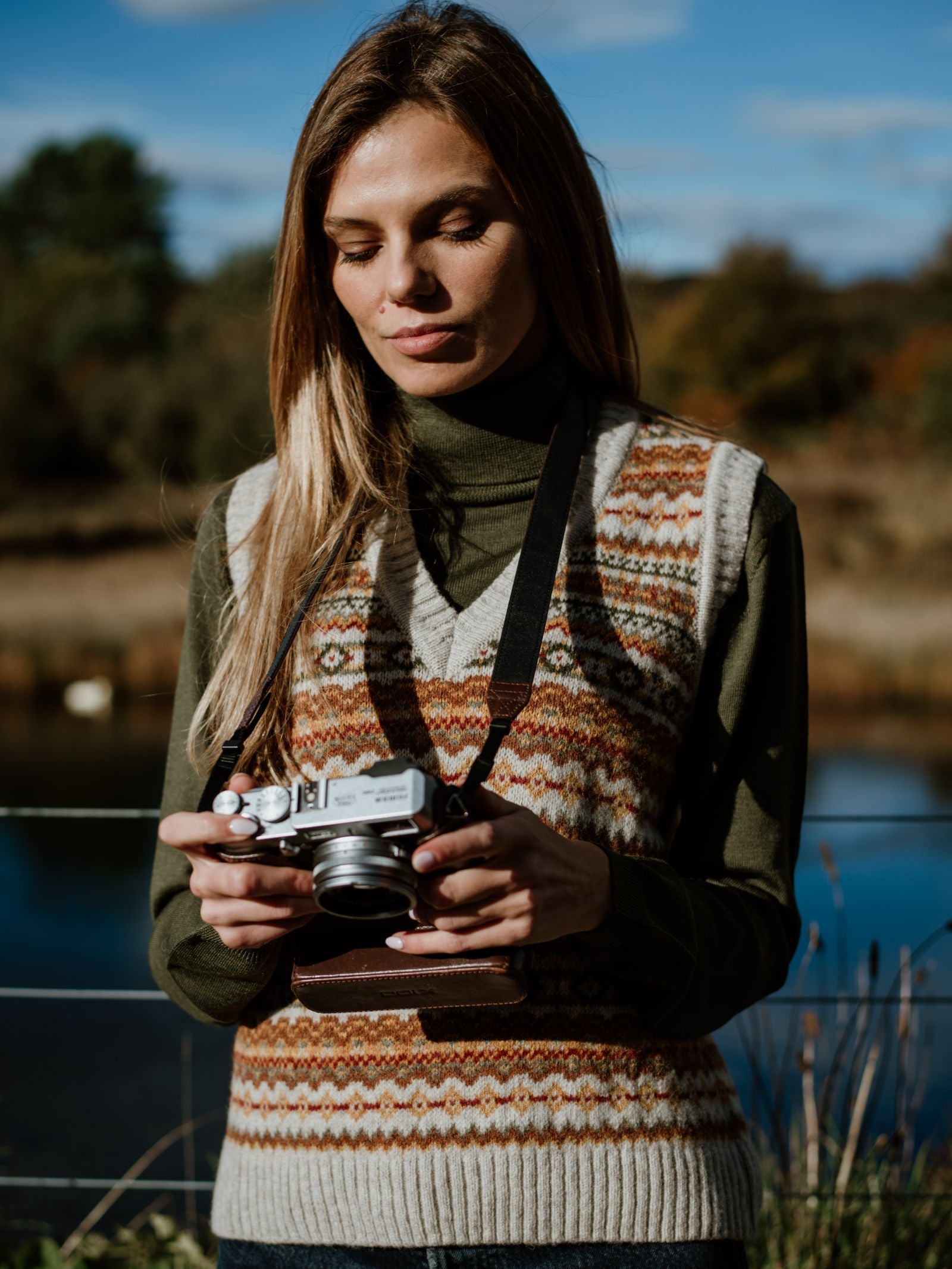 A woman with long blonde hair stands outdoors by a lake, wearing Campbells of Beaulys Natural Fairisle V-Neck Tank over a green turtleneck. Trees and blue sky in the background evoke Scotland’s tranquil beauty as she looks down at her camera.
