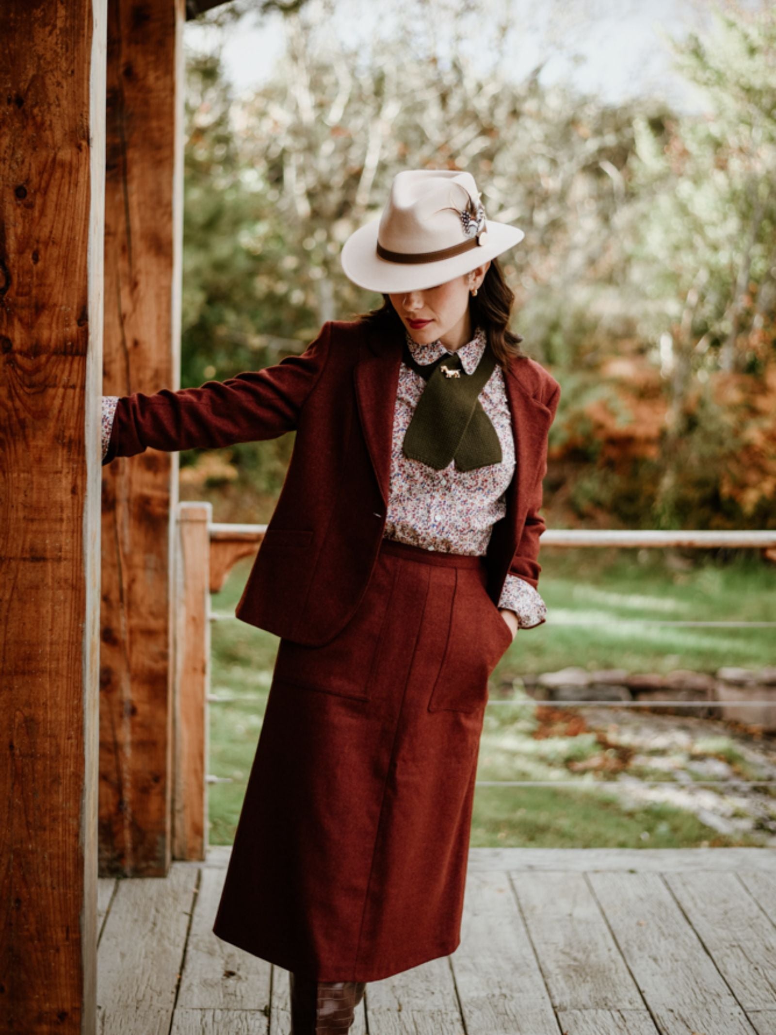 Wearing a Campbell’s of Beauly House Jacket, a woman in a burgundy skirt suit, floral blouse, green necktie, and beige feathered hat stands on a wooden porch amid lush greenery, looking down with one arm resting on the post.