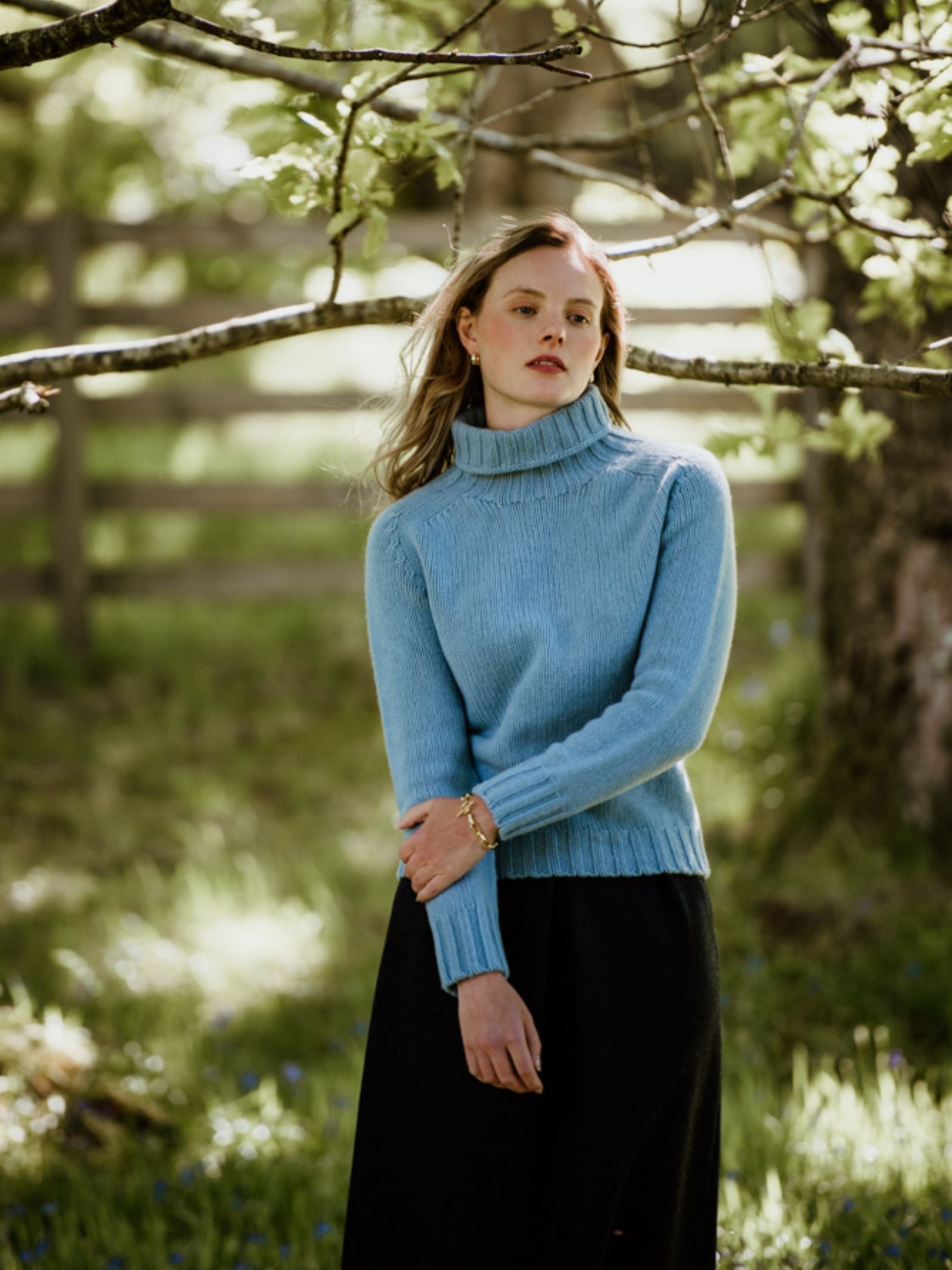 A woman stands outdoors under sunlit tree branches, wearing the Campbells of Beauly Chunky Roll Neck Sweater in light blue and a dark skirt, with a wooden fence blurred in the background.