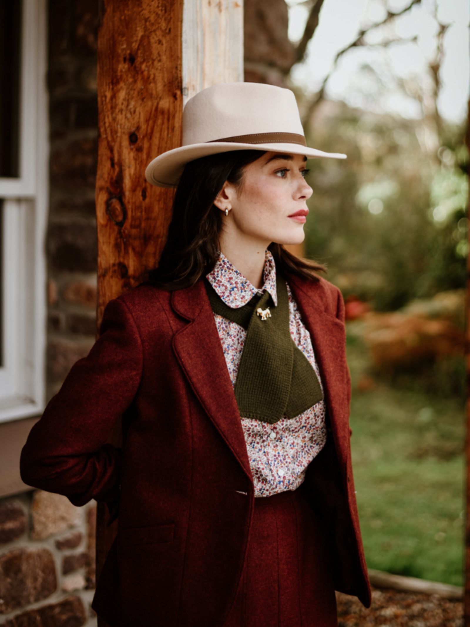 A woman in a maroon suit and floral shirt stands by a wooden post, wearing the Hicks & Brown Gamebird Fedora in light beige. She gazes seriously to the side, framed by lush greenery and trees in the background.