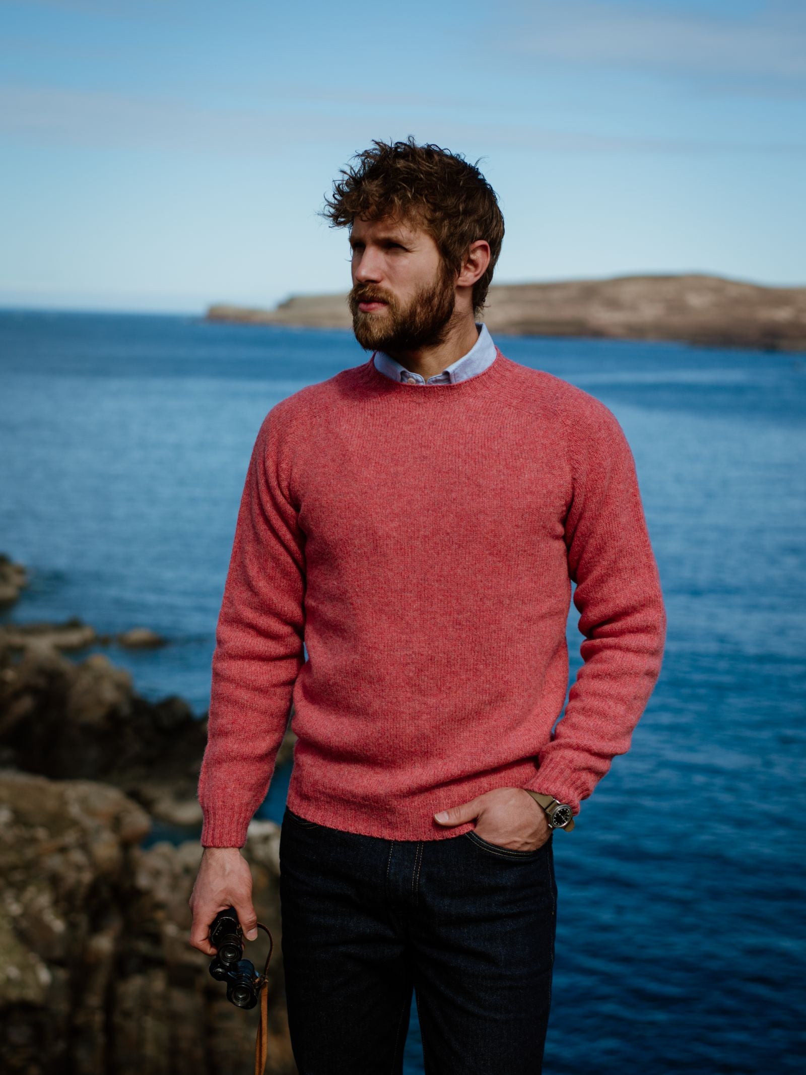 A bearded man in a Campbells of Beauly Shetland Jumper and blue jeans stands on rocky cliffs by the sea, holding binoculars and gazing towards a distant island.