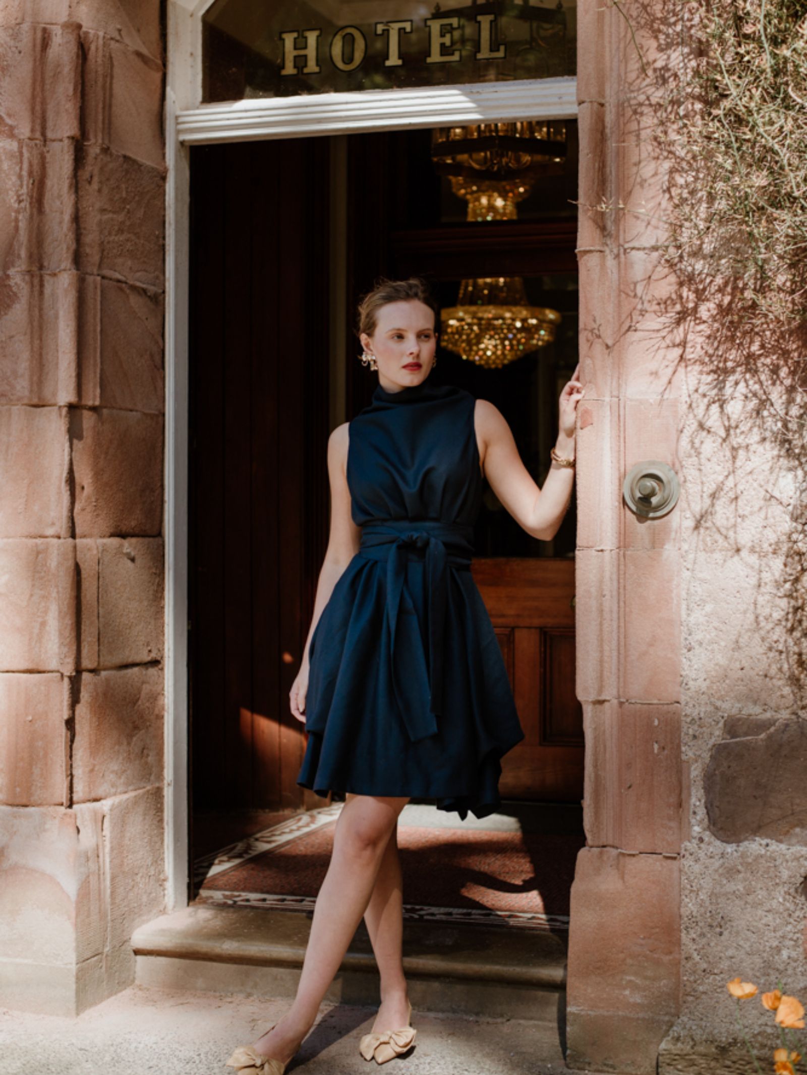 A woman in a sleeveless navy Campbells of Beauly Reversible Wool Wrap Dress and beige flats stands in a stone doorway beneath a HOTEL sign, sunlight flooding the entrance while chandeliers glow inside.