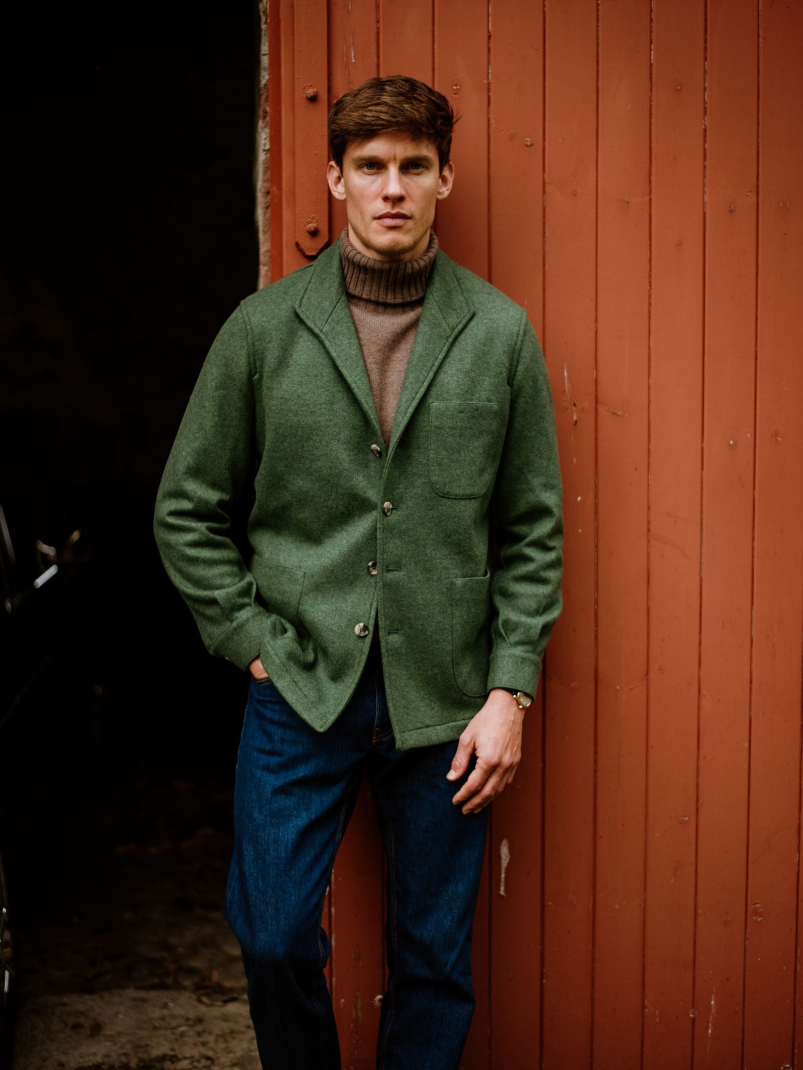 A man wearing an olive Cashmere Teba Jacket by Campbell's of Beauly, a brown turtleneck sweater, and blue jeans stands in front of a reddish-brown wooden wall, looking at the camera with a neutral expression.
