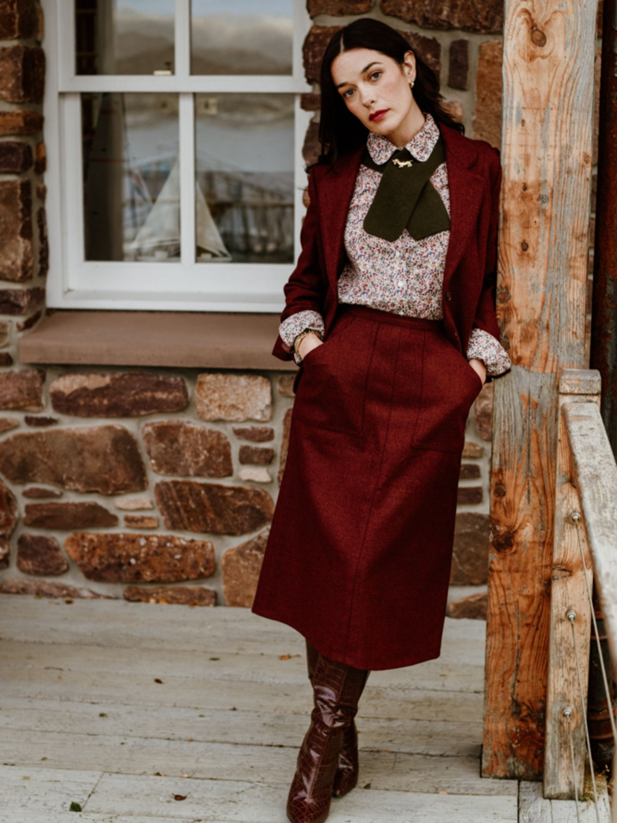 A woman stands on a porch by a stone wall, dressed in a burgundy skirt suit and floral blouse accented with the Campbell’s of Beauly Auld Stock Cashmere Neck Tie—a refined touch that channels classic style and luxury. Brown boots complete the ensemble, while her dark hair and serious expression radiate confidence as she casually slips one hand into her pocket.