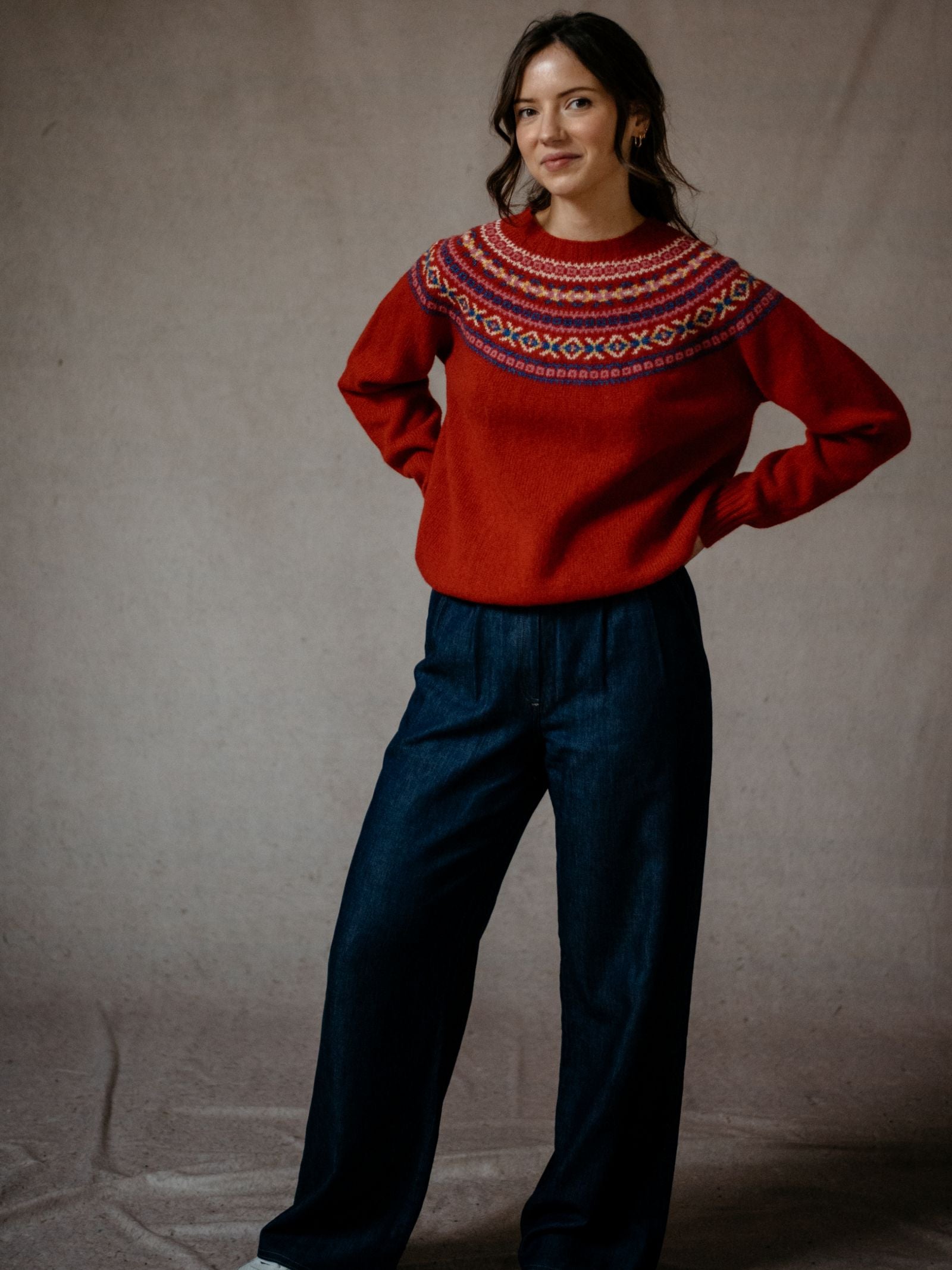 A woman stands against a neutral background, smiling gently with her hands on her hips, wearing Campbells of Beaulys Fairisle Crew Jumper in red with a colorful traditional pattern yoke and dark wide-leg pants.
