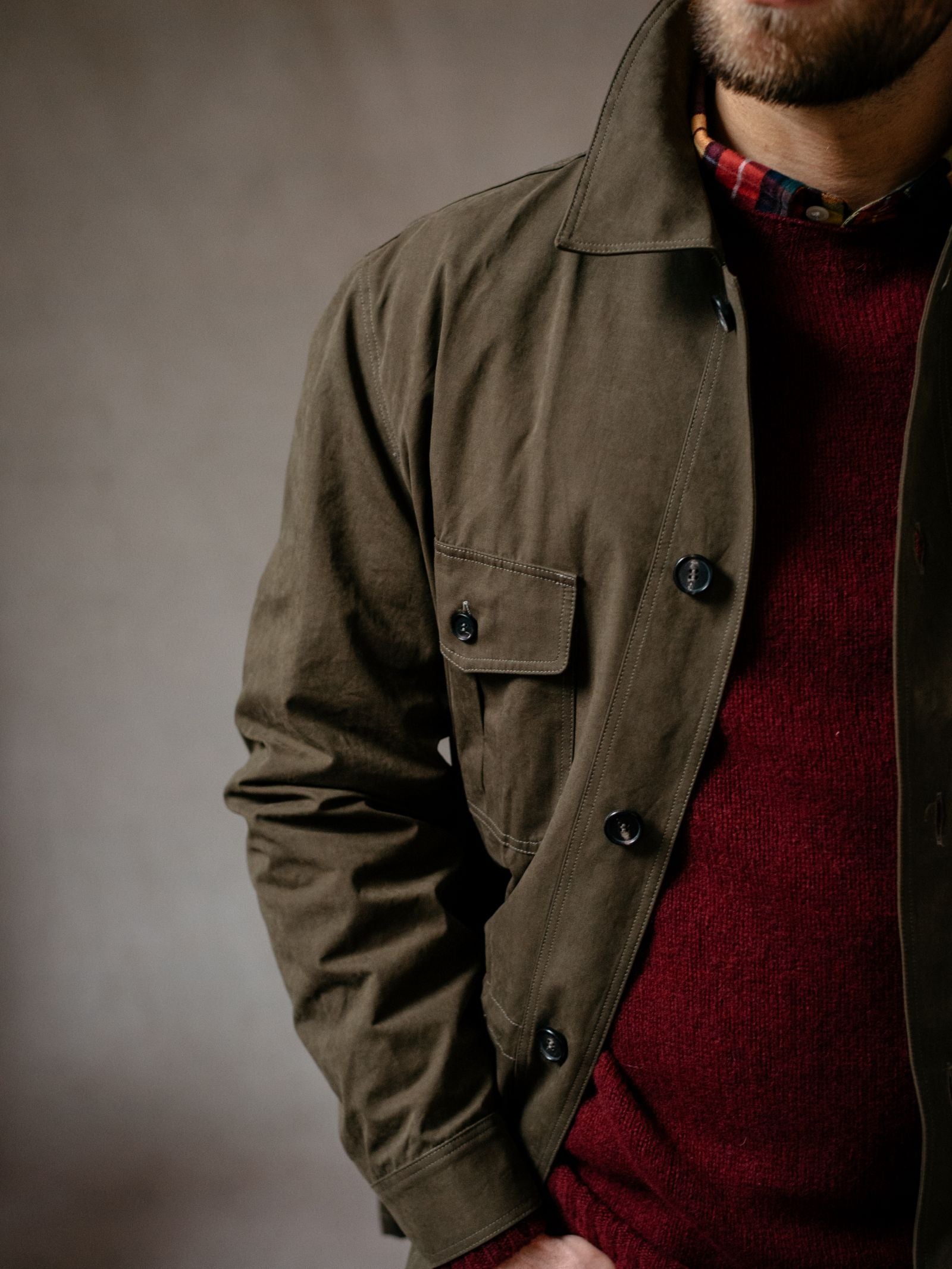 A man wears the Campbells of Beauly Aigas Field Jacket in olive green, made from peach skin fabric, over a red sweater and plaid shirt. He stands with one hand in his pocket against a neutral background, face partially out of frame.