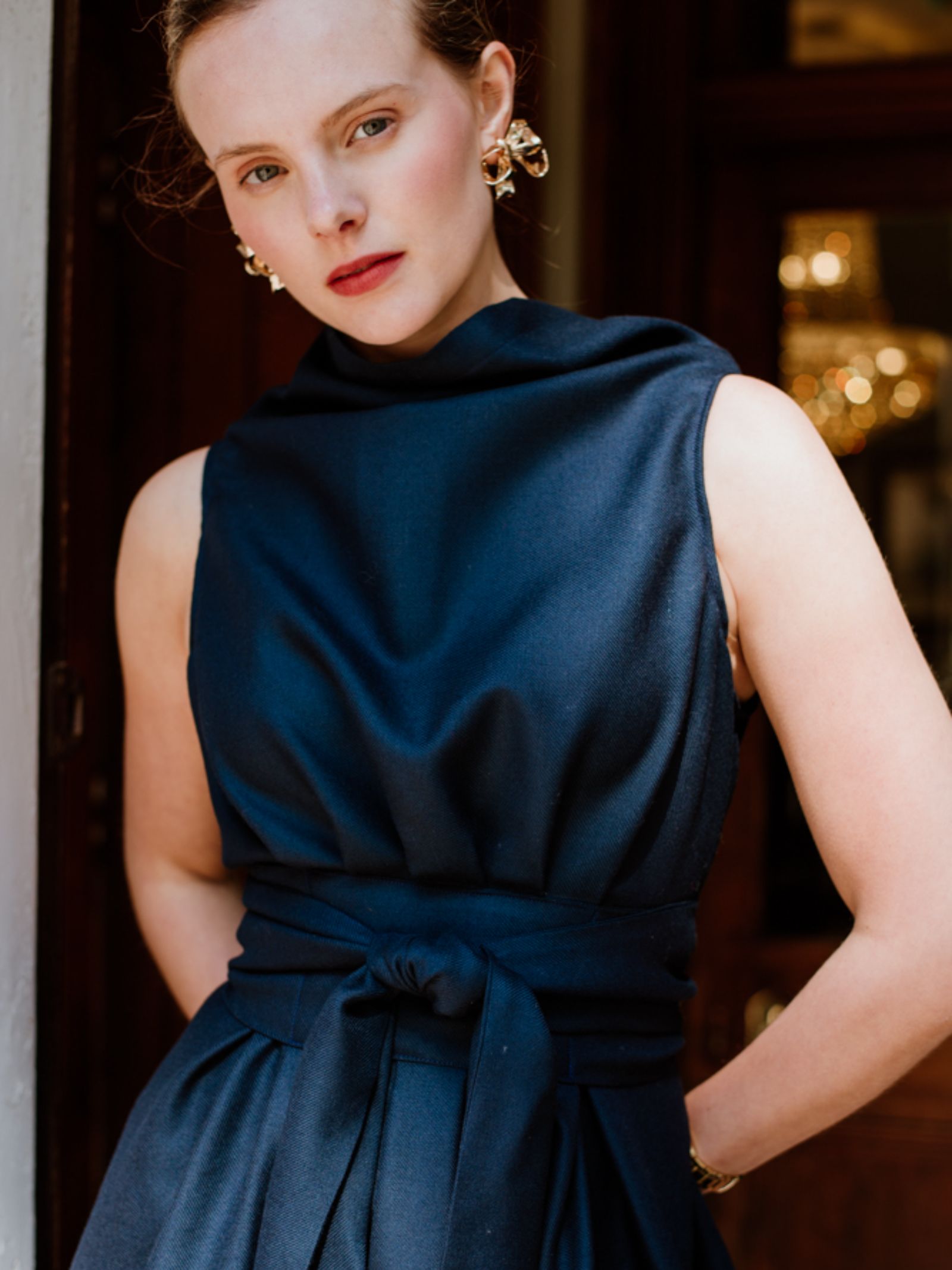 A woman in a sleeveless navy Reversible Wool Wrap Dress by Campbells of Beauly, with a wide sash at the waist, stands indoors under warm light. She faces the camera, wearing gold earrings and red lipstick.