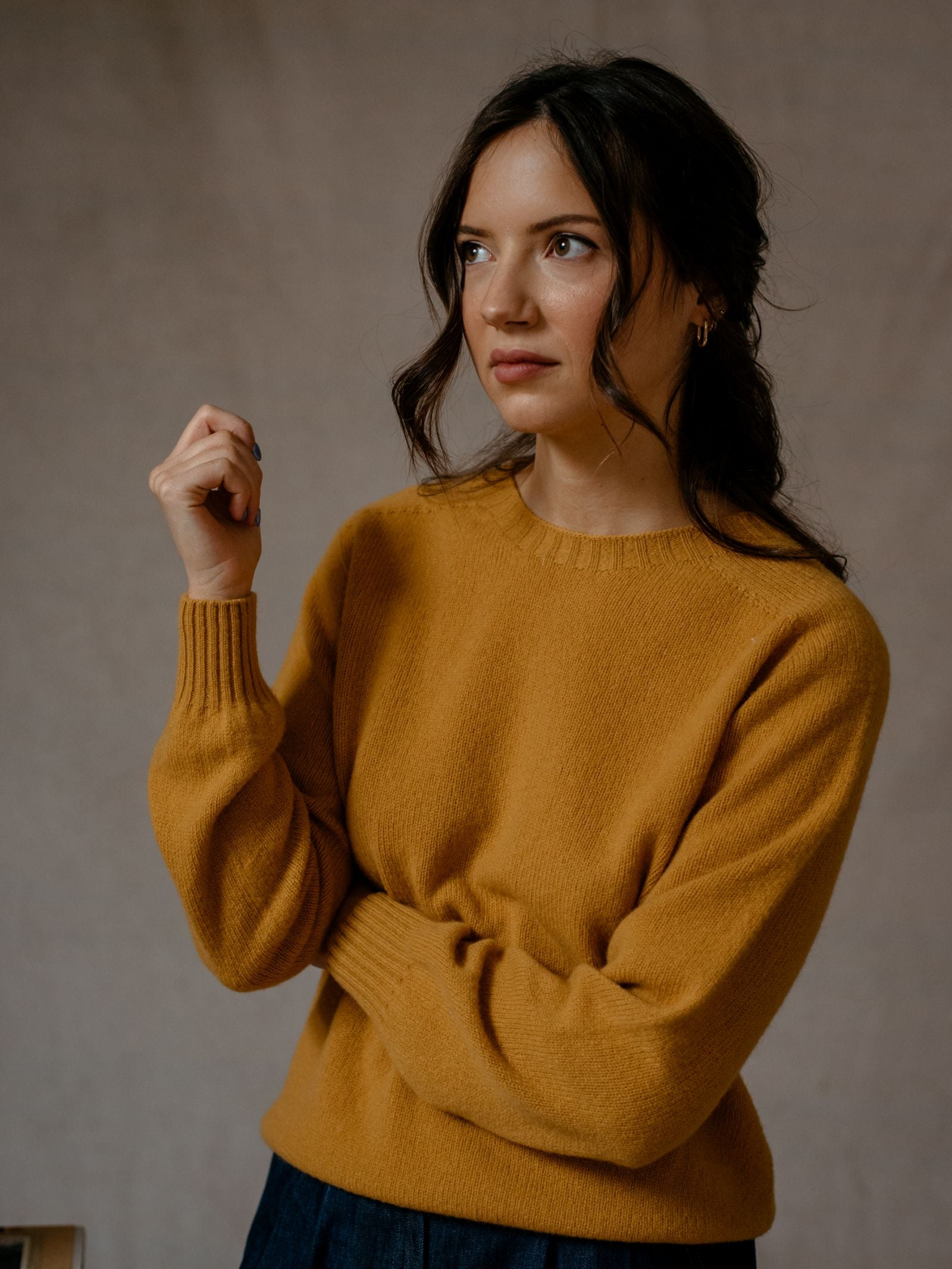 A woman with wavy brown hair, wearing the Campbells of Beauly Geelong Lambswool Crew Neck Jumper in mustard yellow, stands with one arm crossed and looks to the side against a neutral background.