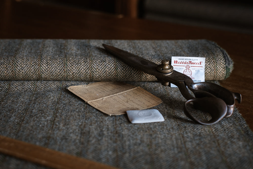 House Tweed 95054 by Campbell’s of Beauly, paired with large vintage scissors, a handwritten note, a piece of chalk, a wooden ruler, and a Harris Tweed label set upon a wooden table, evokes the timeless tradition of bespoke tailoring. The classic herringbone pattern of this jacketing cloth further highlights the enduring craftsmanship associated with House Tweed 95054.