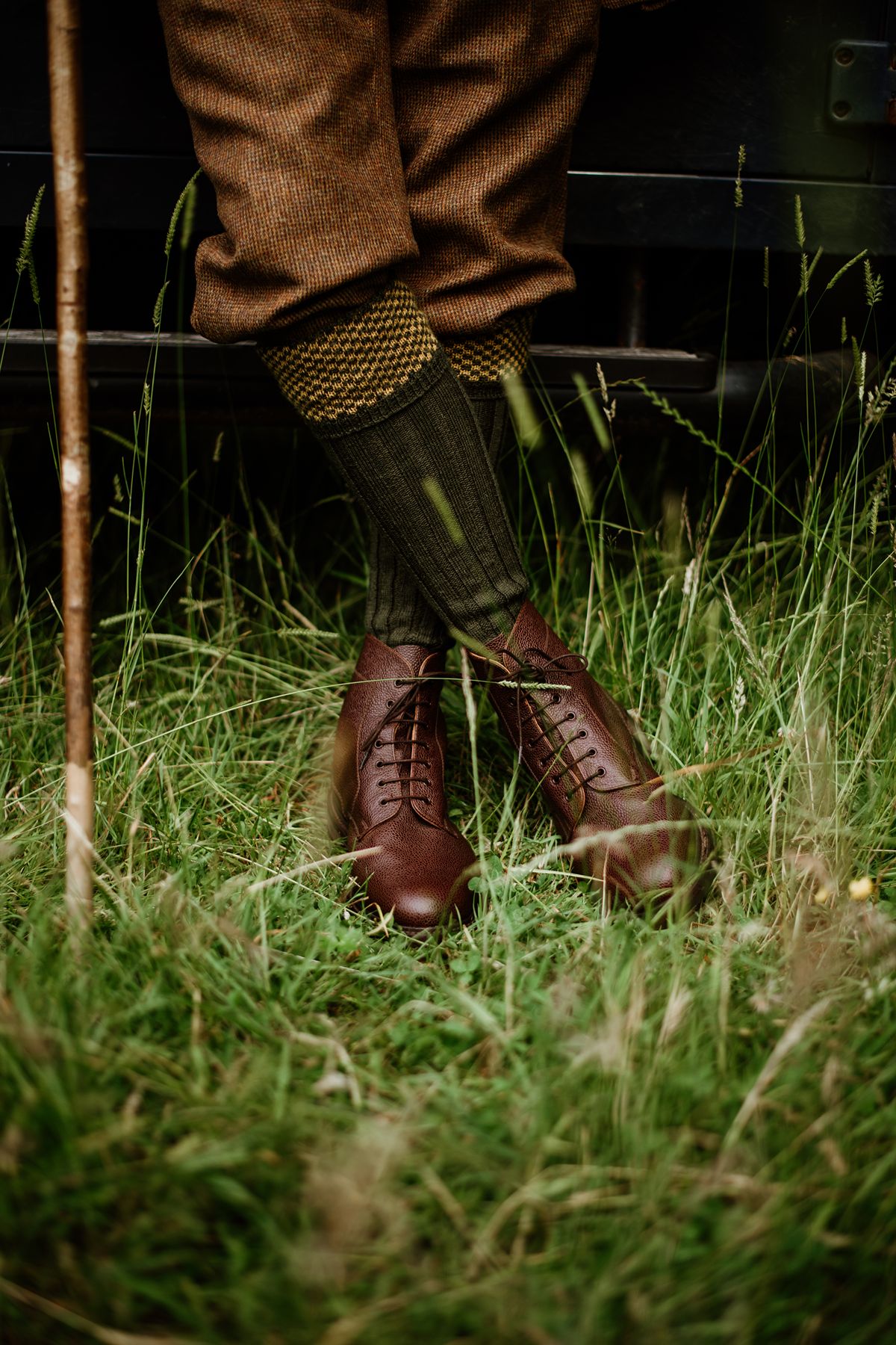 A person wearing brown trousers, patterned socks, and lace-up brown boots standing in tall grass.