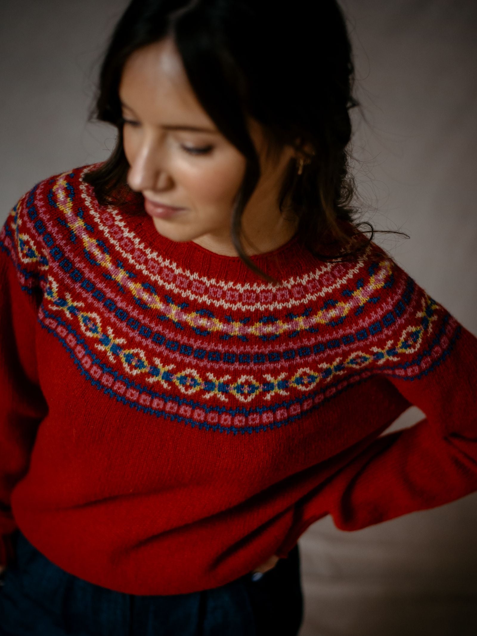 A woman with wavy brown hair wears the Campbells of Beauly Fairisle Crew Jumper, made from superfine Geelong lambswool, featuring a colorful traditional pattern on the yoke. She gazes downward against a soft, neutral background.