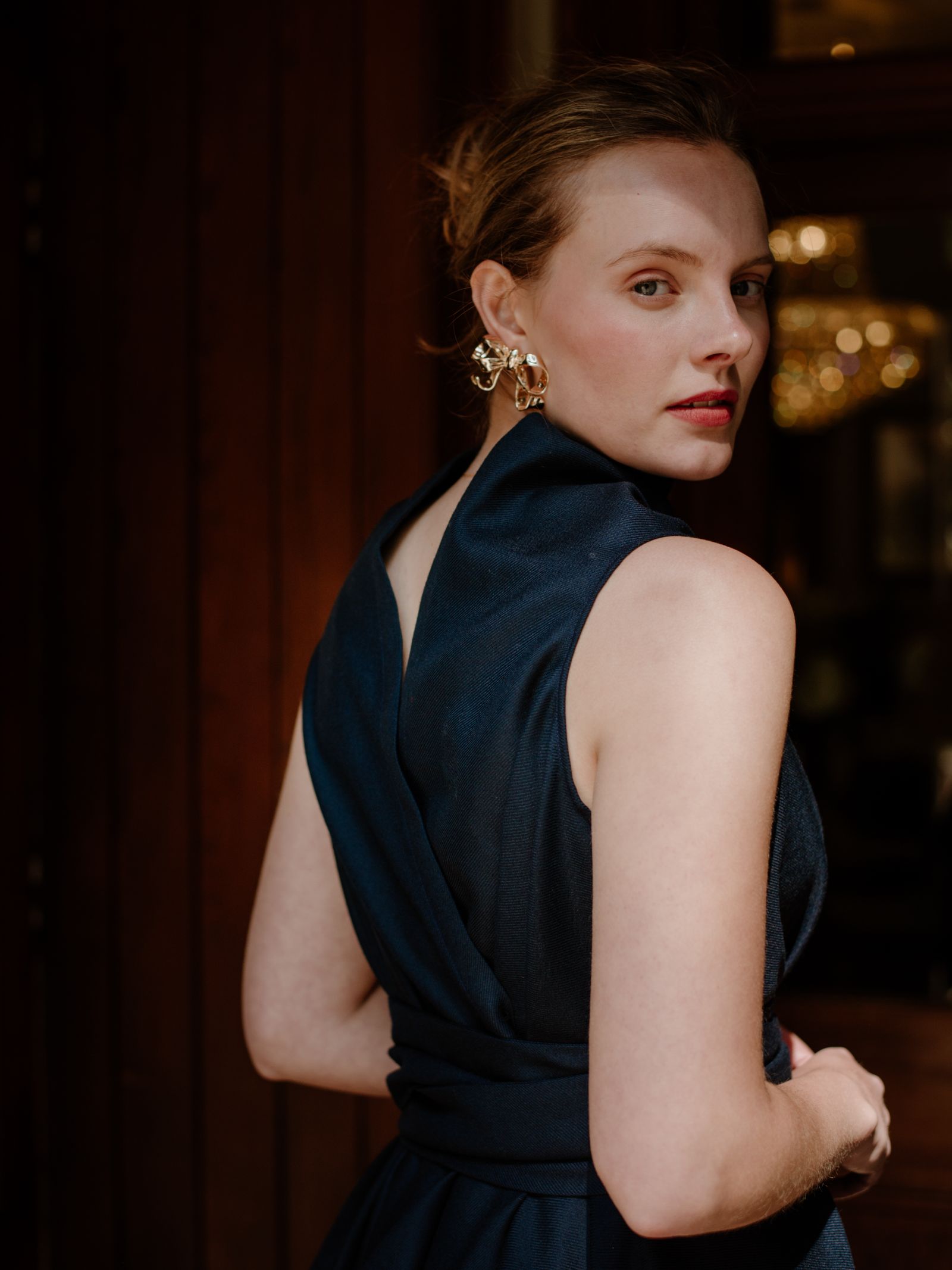 A woman with light skin and light brown hair in an updo wears a sleeveless navy Reversible Wool Wrap Dress by Campbells of Beauly and gold earrings, standing sideways in warm light with a chandelier and dark wood wall behind her.