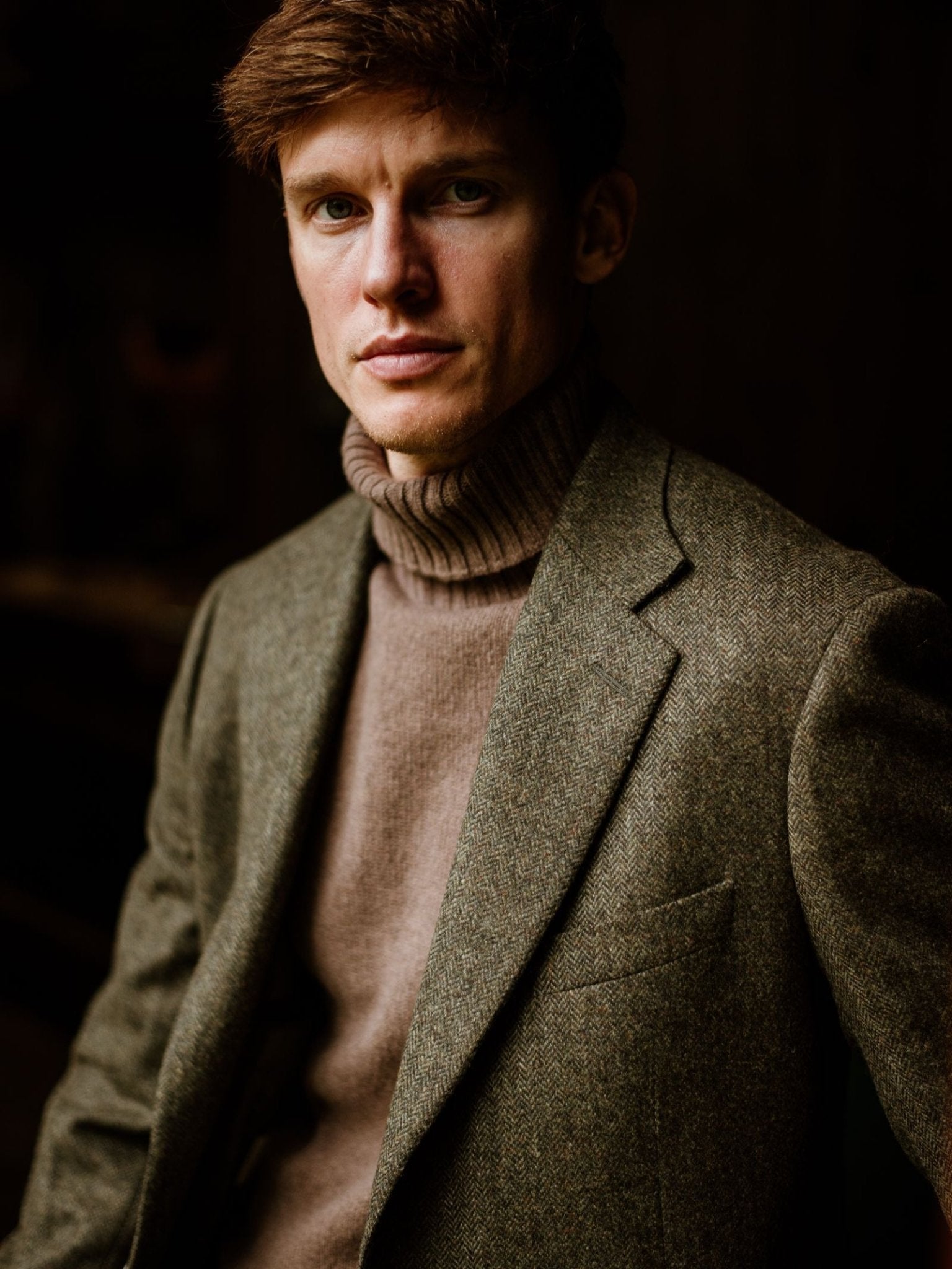 A young man with short brown hair wears the Campbell's of Beauly Auld Stock Lambswool Roll Neck and a green tweed blazer, looking directly at the camera against a dark background.