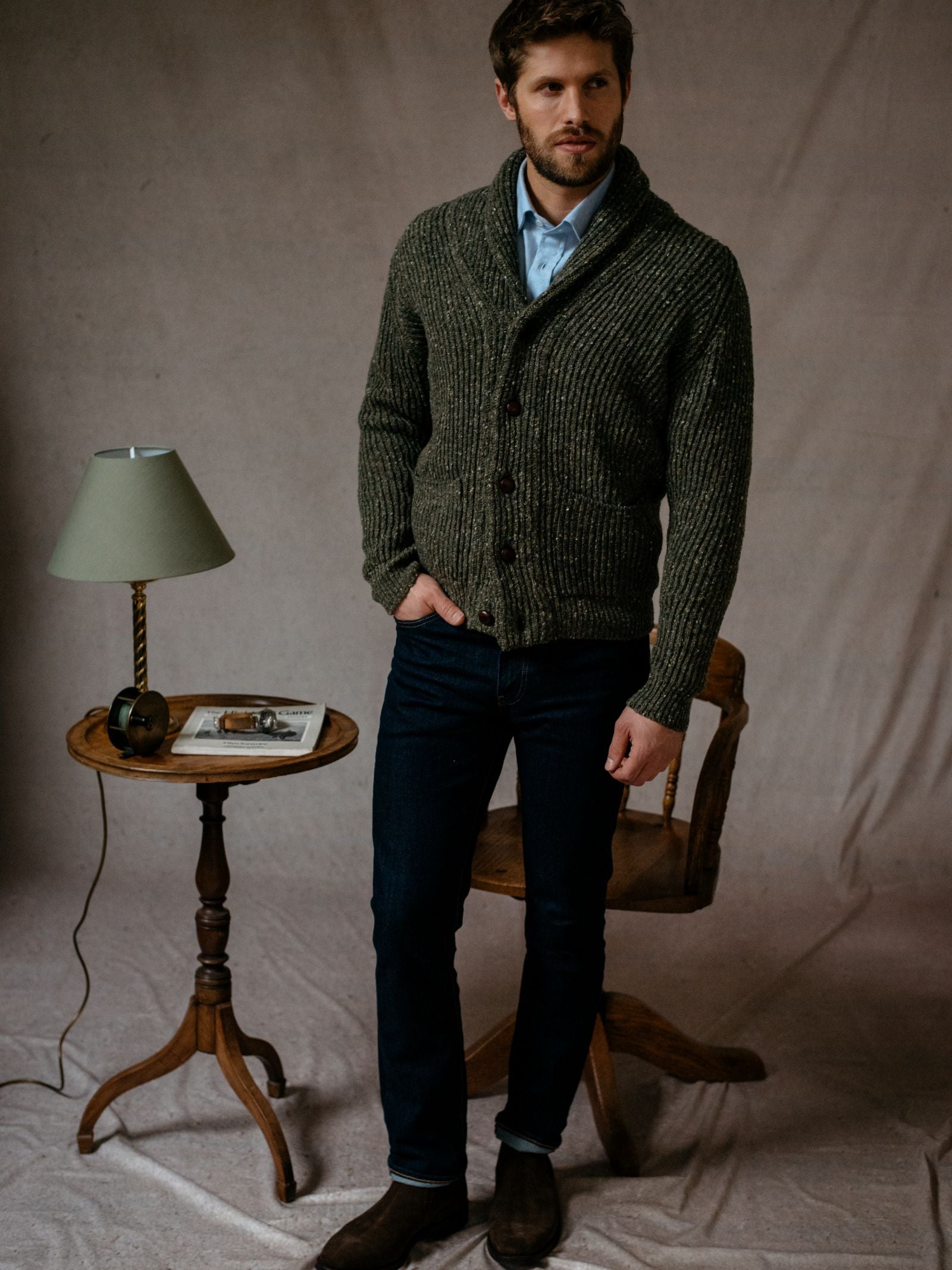 A man stands indoors on a draped cloth, wearing the Campbells of Beauly Donegal Lambswool Shawl Collar Cardigan with leather buttons, blue shirt, dark jeans, and brown boots. Beside him is a small round table and wooden chair.
