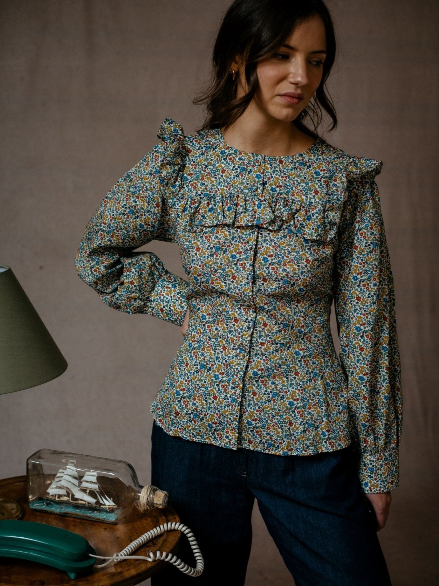 A woman in a Campbells of Beauly Liberty Print Peplum Shirt and dark jeans stands indoors by a table with a green lamp, vintage phone, and ship in a bottle, looking down with one hand on her hip.