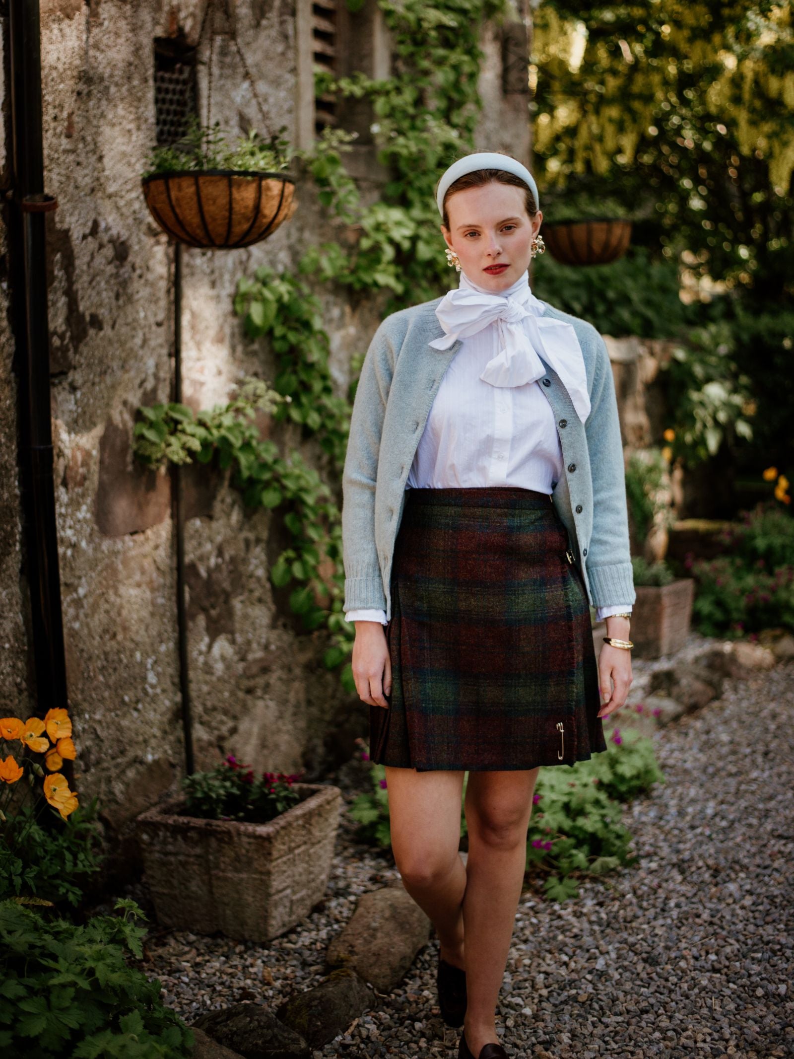 A woman in a white blouse with a bow, light blue cardigan, and Campbells of Beauly Tweed Kilt stands on a gravel path by a stone wall decorated with hanging baskets and green plants, looking at the camera.