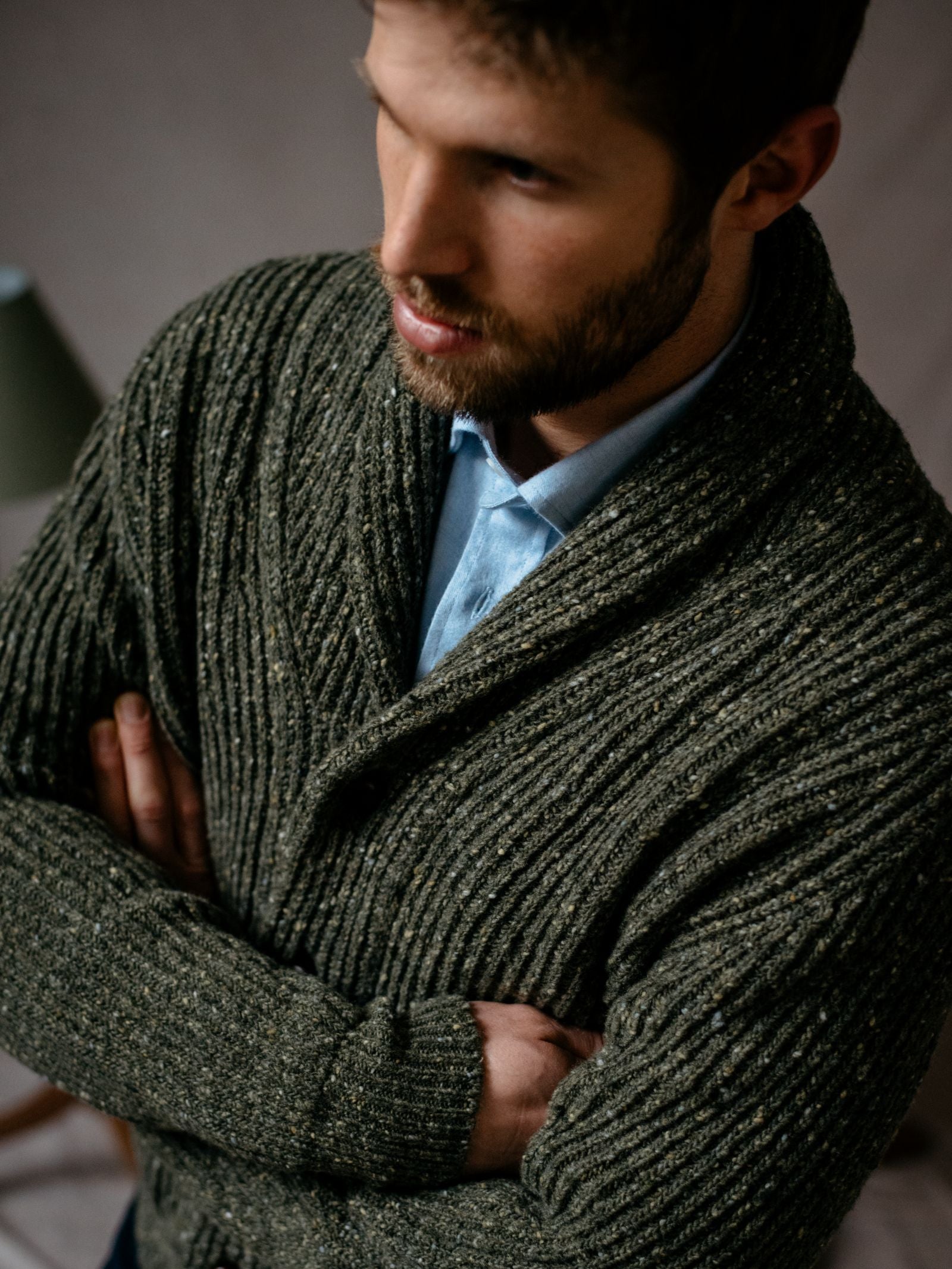 A bearded man with short hair stands with arms crossed, wearing Campbells of Beauly Donegal Lambswool Shawl Collar Cardigan in dark green over a light blue shirt, gazing slightly down and to the side against a softly blurred background.